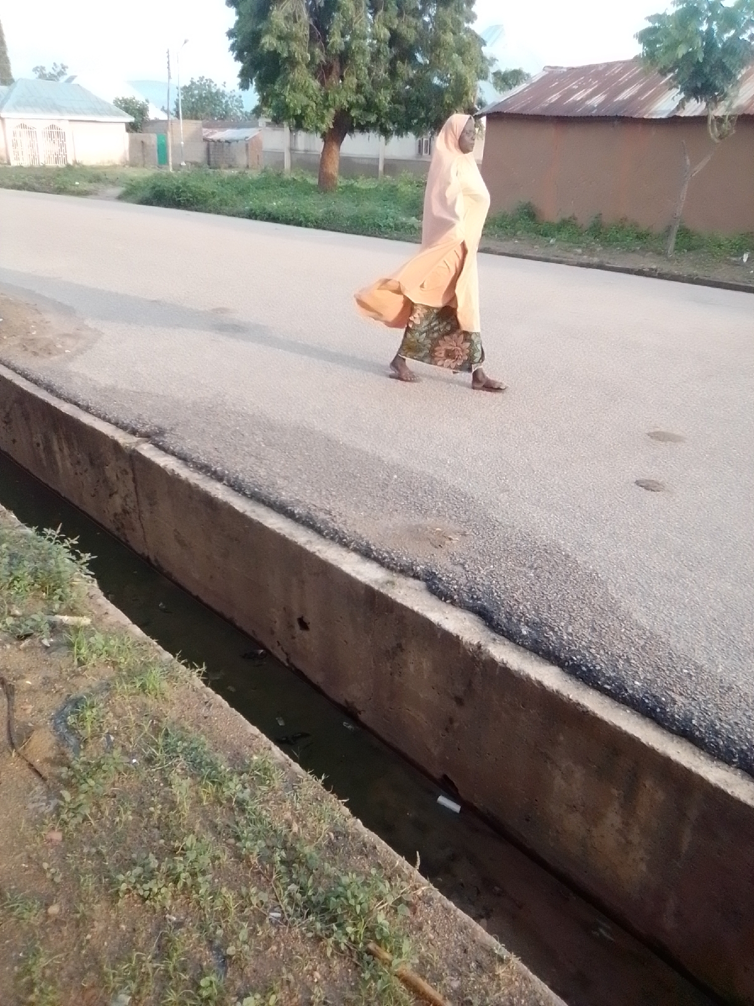 The image depicts a scene in the city of Gombe, Nigeria, near the Federal College of Education. A woman, dressed in a light orange hijab and a patterned skirt, walks along the side of a paved road. She is seen from the side, moving towards the right of the frame. The road has a noticeable concrete drainage channel running alongside it, with some murky water visible within. Vegetation, including grass and small plants, grows along the edge of the drainage channel. In the background, there are buildings with corrugated iron roofs, trees, and a street lamp. The lighting suggests it is either early morning or late afternoon.