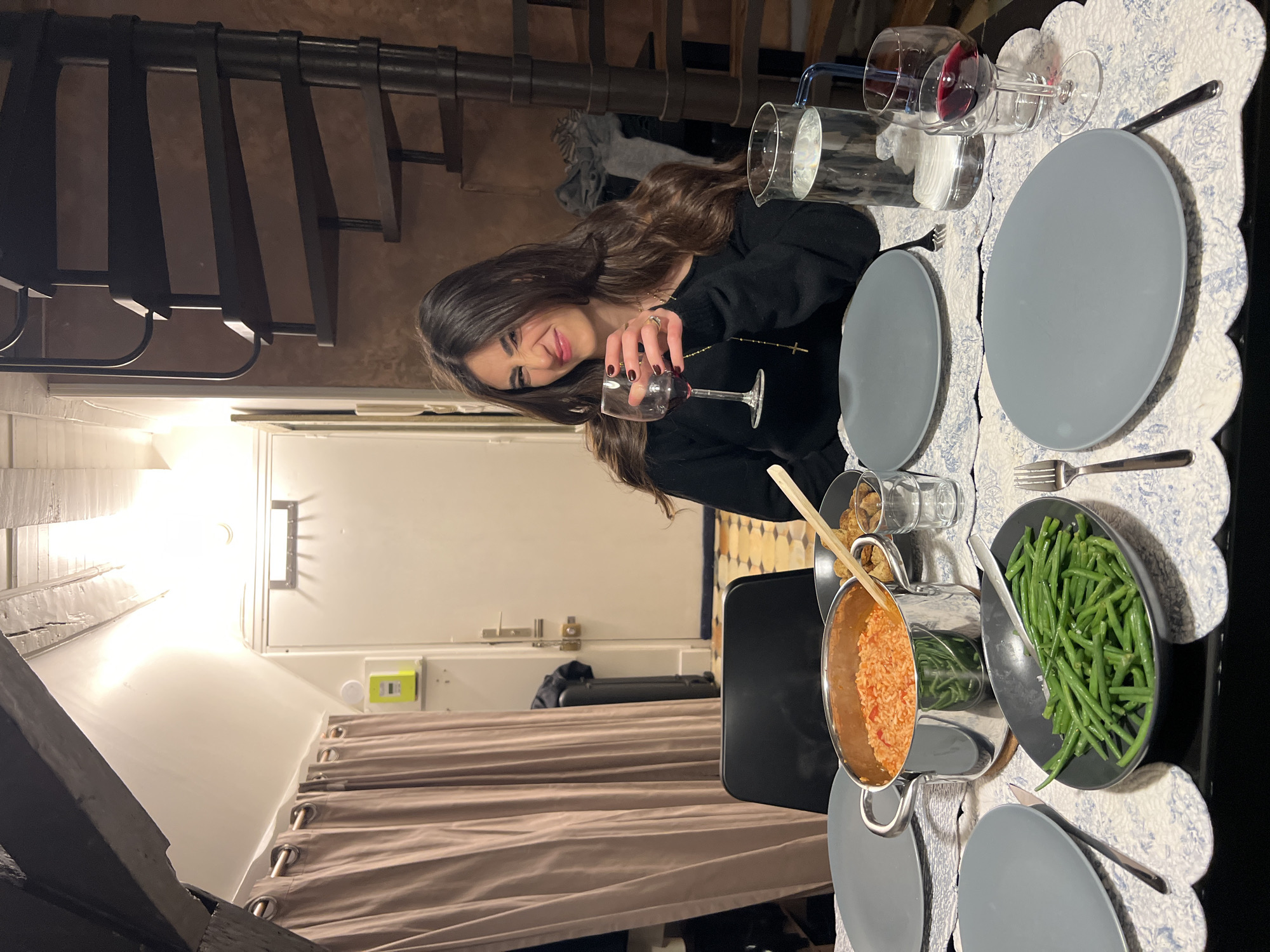 An indoor scene depicts a young woman dining at a table, set for at least two people. The image is taken from a slightly elevated perspective, looking down at the table and the woman.
The central figure is a young woman with long, dark hair, wearing a black long-sleeved top and a gold cross necklace. She is smiling broadly and looking directly at the camera while holding a wine glass with a small amount of red wine. Her left hand, adorned with a ring, is visible.
The dining table is covered with a white and blue scalloped placemat. On the table, there are two grey dinner plates, two forks, and a set of wine glasses, one of which contains red wine. A glass carafe filled with water is also present. Food items include a metallic pot holding what appears to be orange-colored rice or risotto, a black bowl filled with green beans, and a small glass bowl containing small, fried items. A wooden spoon rests in the pot of rice.
In the background, a dark spiral staircase is prominently featured on the left, winding upwards. To the right of the staircase, a brightly lit wall section reveals a white door with a handle and latch. Below this, a light brown curtain is pulled back, revealing a glimpse of an interior space. Artificial lighting illuminates the scene, suggesting it is evening or night. The overall atmosphere appears relaxed and convivial. No discernible text is visible in the image.