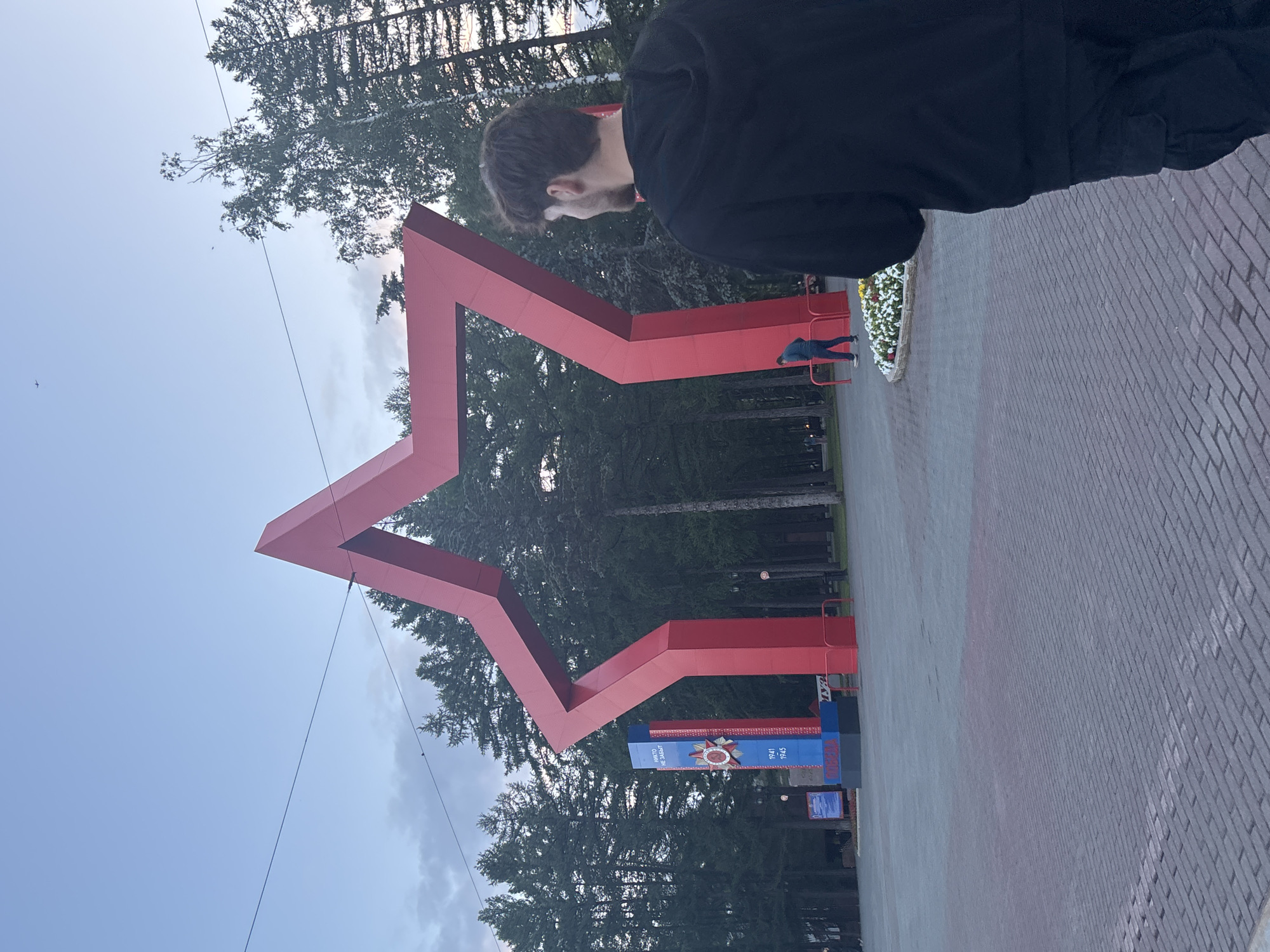 The image depicts a view of the Eternal Flame memorial complex in Ufa, Russia. In the foreground, a young man with dark, curly hair is seen from behind, wearing a black hoodie and black cargo pants. He is facing away from the camera, looking towards a large, red, five-pointed star structure that dominates the scene. The star structure is a prominent monument, likely part of the memorial.
To the left of the star, a blue and red monument with an emblem and text in Russian is visible. The text on the blue part reads "НИКТО НЕ ЗАБЫТ" (No one is forgotten), and below it are the years "1941" and "1945". The red part has the word "ПОБЕДА" (Victory) written on it.
In the background, a forest of evergreen trees forms a dense backdrop. Several streetlights are visible among the trees. A person in a blue shirt and dark pants is walking near the red star. The ground in the foreground is paved with grey bricks. The sky is a pale blue with some clouds, and there are a couple of small planes or drones visible in the distance. The overall lighting suggests it is either early morning or late evening.