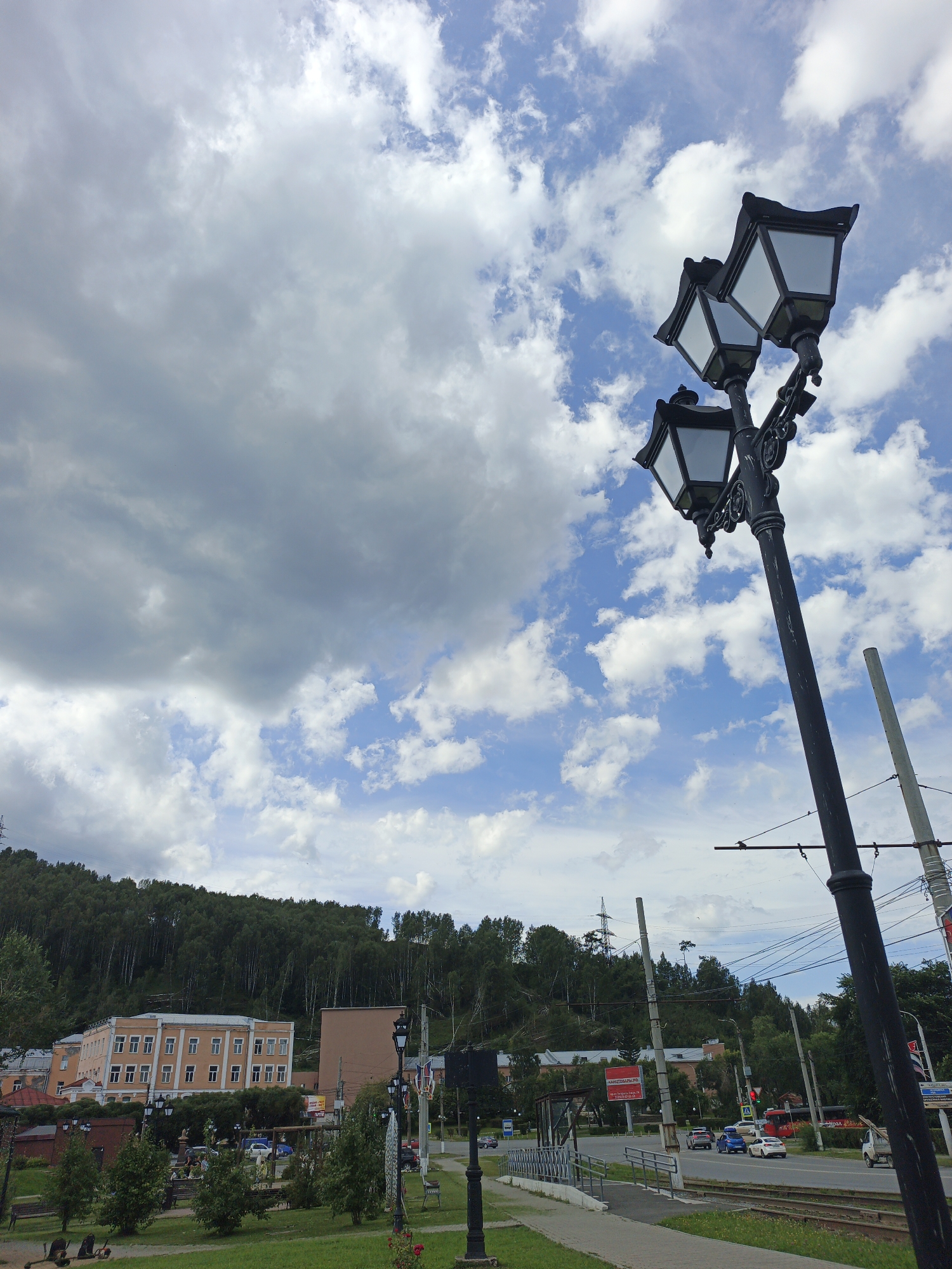 This is an outdoor, eye-level, full shot photograph taken on a cloudy day in the city of Zlatoust, Russia. In the foreground, a black, ornate lamppost with three lamps is positioned to the right of the frame, angled upwards. Behind it, a paved walkway runs along the right side of the image, bordered by a grassy area and trees. A set of trolley tracks runs parallel to the walkway. In the mid-ground, a street with several cars and a red trolley is visible. Beyond the street, buildings with light-colored facades and rows of windows are situated at the base of a densely wooded hill. The hill, covered in green trees, dominates the background and extends across the upper portion of the image. The sky above is a mix of blue patches and large, white and grey clouds.