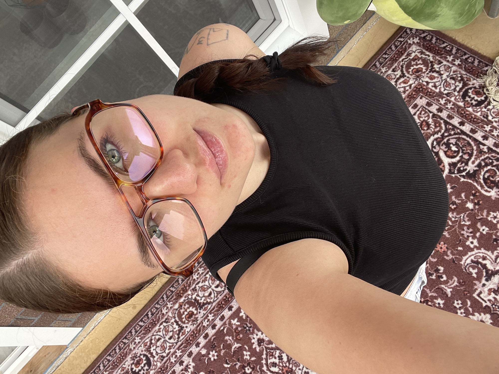 A selfie of a woman with green eyes and glasses wearing a black top. Her hair is brown and pulled back with a black hair tie. She has a tattoo on her arm. The background includes a patterned rug and what appears to be a window or door. It is impossible to determine the location with the given information.