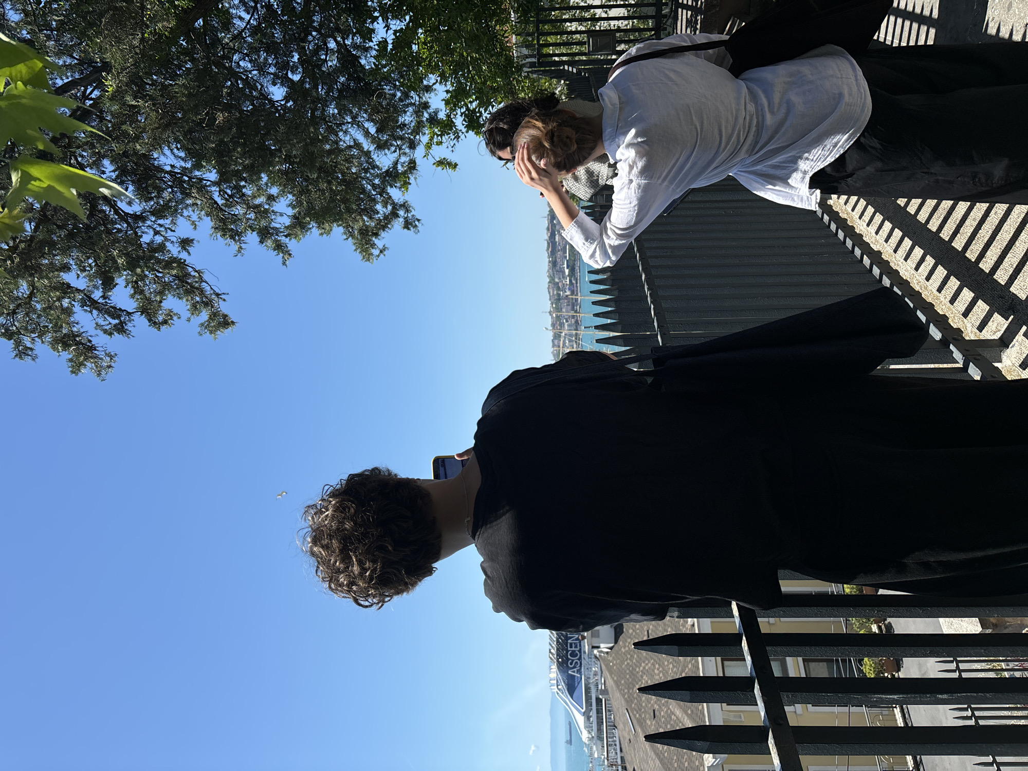 Two people are looking out over a railing at a view of the city of Beyoğlu, Turkey. One person wears a white, long-sleeved shirt and black pants, and has their hand to their forehead. The second person wears a black long-sleeved shirt and looks down at a mobile phone. The railing is black metal. There are trees visible in the upper left of the image and a blue sky in the center left.