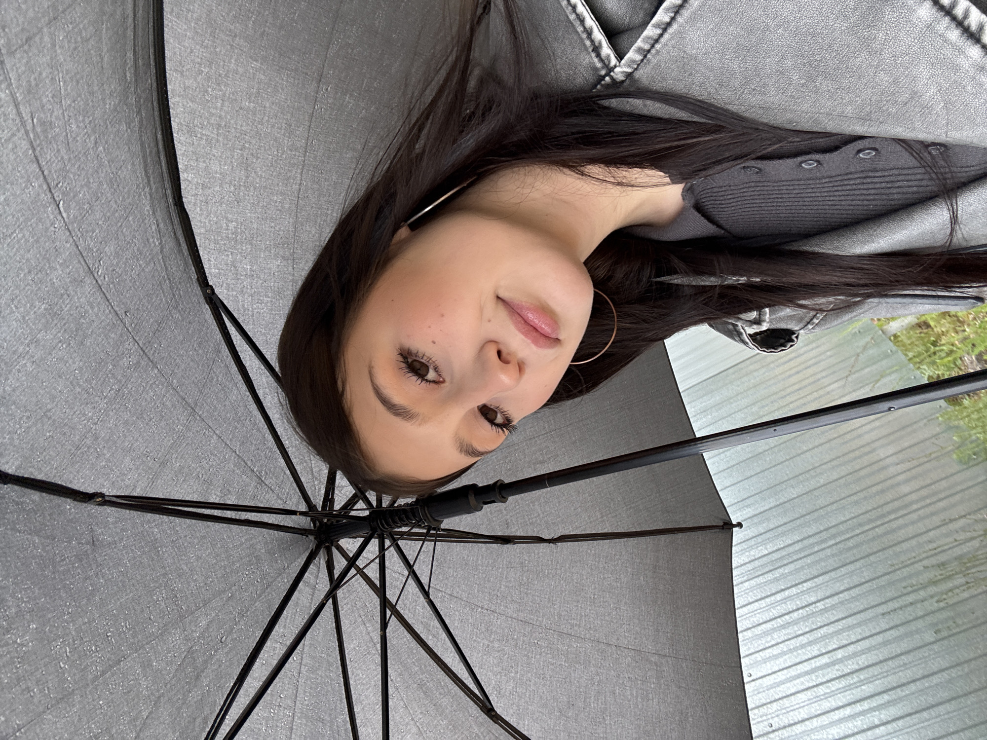 A young woman with long, dark hair is captured in a close-up portrait, looking directly at the camera from beneath a grey umbrella. The umbrella's canopy shows numerous small water droplets, indicating recent or ongoing rain. The woman has prominent eyelashes, defined eyebrows, and light-colored lips. She wears large hoop earrings and is dressed in a dark grey ribbed top or cardigan underneath a lighter grey jacket, possibly leather or faux leather, with visible stitching. In the background, beyond the umbrella and the subject, there are blurred elements including a corrugated metal surface, possibly part of a building or fence, and some indistinct green foliage. The lighting suggests it is daytime, and the overall scene conveys an outdoor setting in Фоки, Russia, likely during or after rainy weather. There is no visible text in the image.