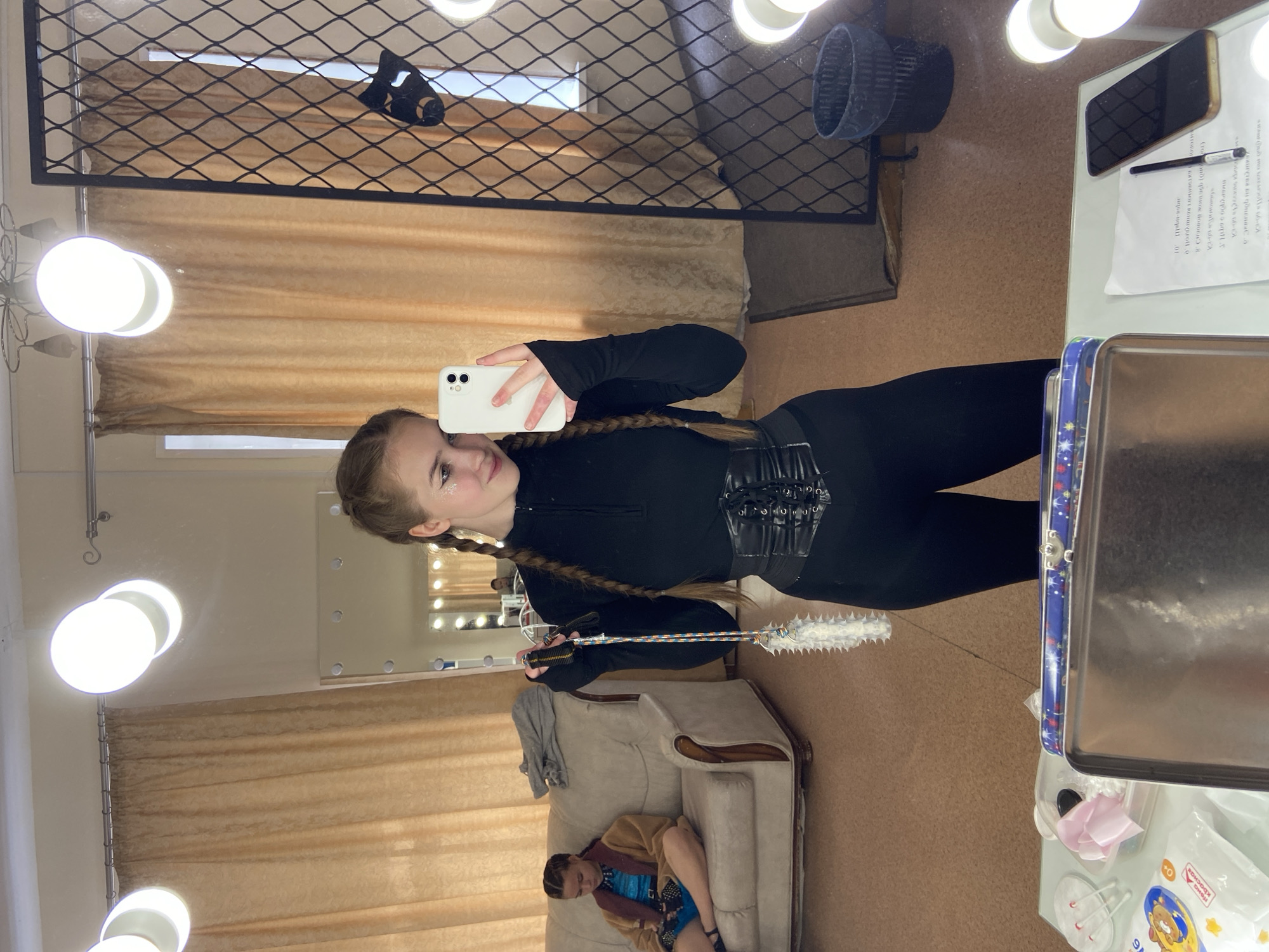 The image captures a young woman taking a mirror selfie in what appears to be a backstage or dressing room area. She is wearing a black, form-fitting outfit with a corset-like belt and has her hair in two braids. In her hand, she holds a fluffy object attached to a colorful string. To her left, another young woman is seated on a couch, wrapped in a brown robe, looking down at something in her lap. The room features tan curtains, a large mirror with vanity lights, and various items on a table in the foreground, including a metal box, makeup supplies, a phone, and a piece of paper with text. The overall environment suggests a preparation area for a performance or event. The location is identified as Chaikovsky, Russia.