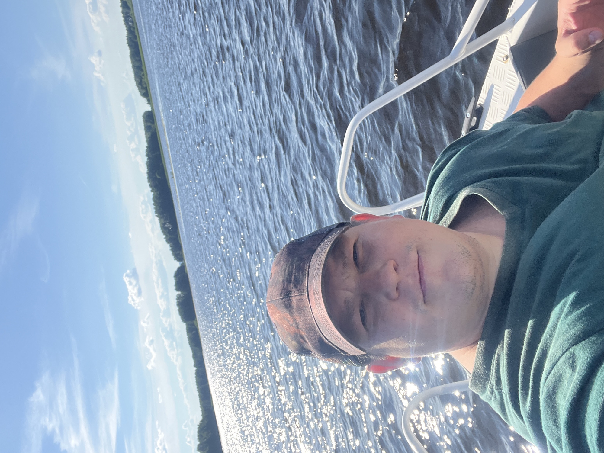 The image shows a person taking a selfie while on a boat. The person is wearing a camouflage baseball cap and a green t-shirt. They have a slight smile on their face. The boat appears to be on a body of water, possibly a lake or river, with trees lining the shore in the background. The water is rippling and reflecting the sunlight, creating a sparkling effect. The sky is blue with some scattered clouds. The sun appears to be high in the sky, suggesting it is daytime, likely late morning or early afternoon. The overall mood seems relaxed and enjoyable, indicating the person is having a pleasant time on the water. There is no visible text in the image. The location context provided is Сюктерка, Russia.