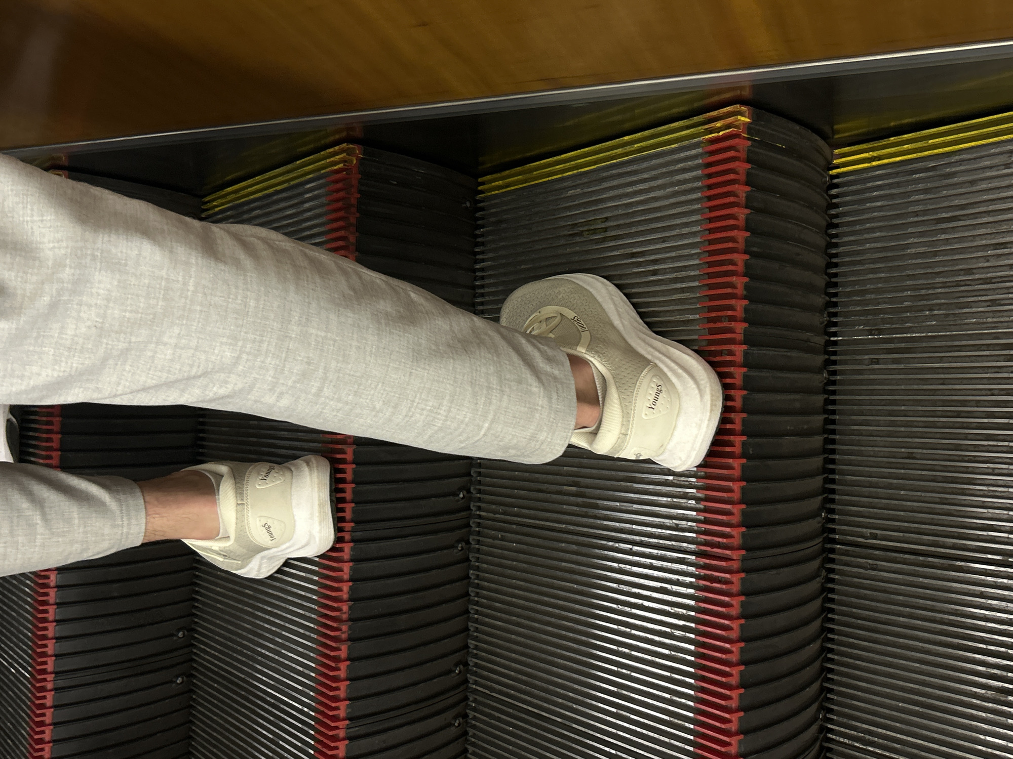 The image shows a person's legs and feet on an escalator. The person is wearing light grey trousers and cream-colored sneakers with the brand name "Youngs" visible on the side of the shoe. The escalator steps are dark grey with red and yellow warning strips. The background is a dark, metallic surface, possibly part of the escalator's casing or the surrounding structure. The lighting suggests an indoor environment, possibly a public building or transportation hub. The scene is captured from a low-angle perspective, focusing on the feet and lower legs. There are no clear indications of time of day or weather conditions. The overall impression is that of a mundane moment captured in an urban setting in Saint Petersburg, Russia.