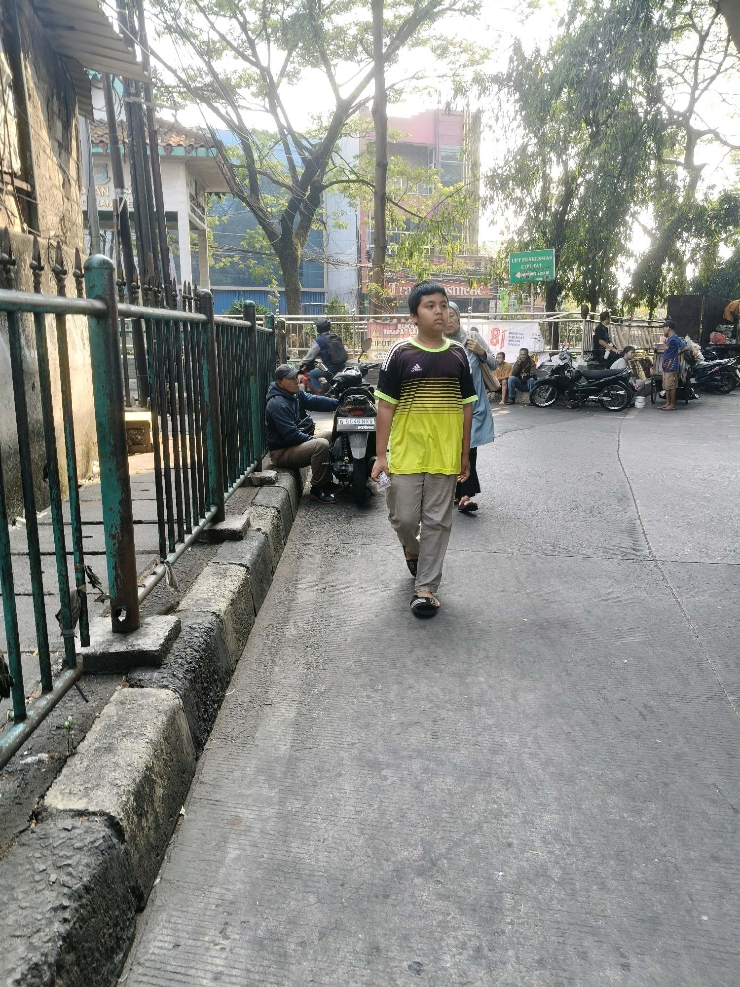 The image depicts a street scene in South Tangerang, Indonesia, under bright daylight. In the foreground, a young boy walks towards the viewer, wearing a yellow and black striped t-shirt and light-colored pants. He is holding a piece of paper in his right hand. Behind him, other people are visible, including a woman in a headscarf. Several motorcycles are parked or being ridden in the background. A man in a dark jacket and cap sits on the curb next to a motorcycle. The environment includes a sidewalk with a metal fence, trees with lush foliage, and buildings in the distance. One building has a green sign that reads "UPT PUSKESMAS CIPELAT". The street surface is asphalt.