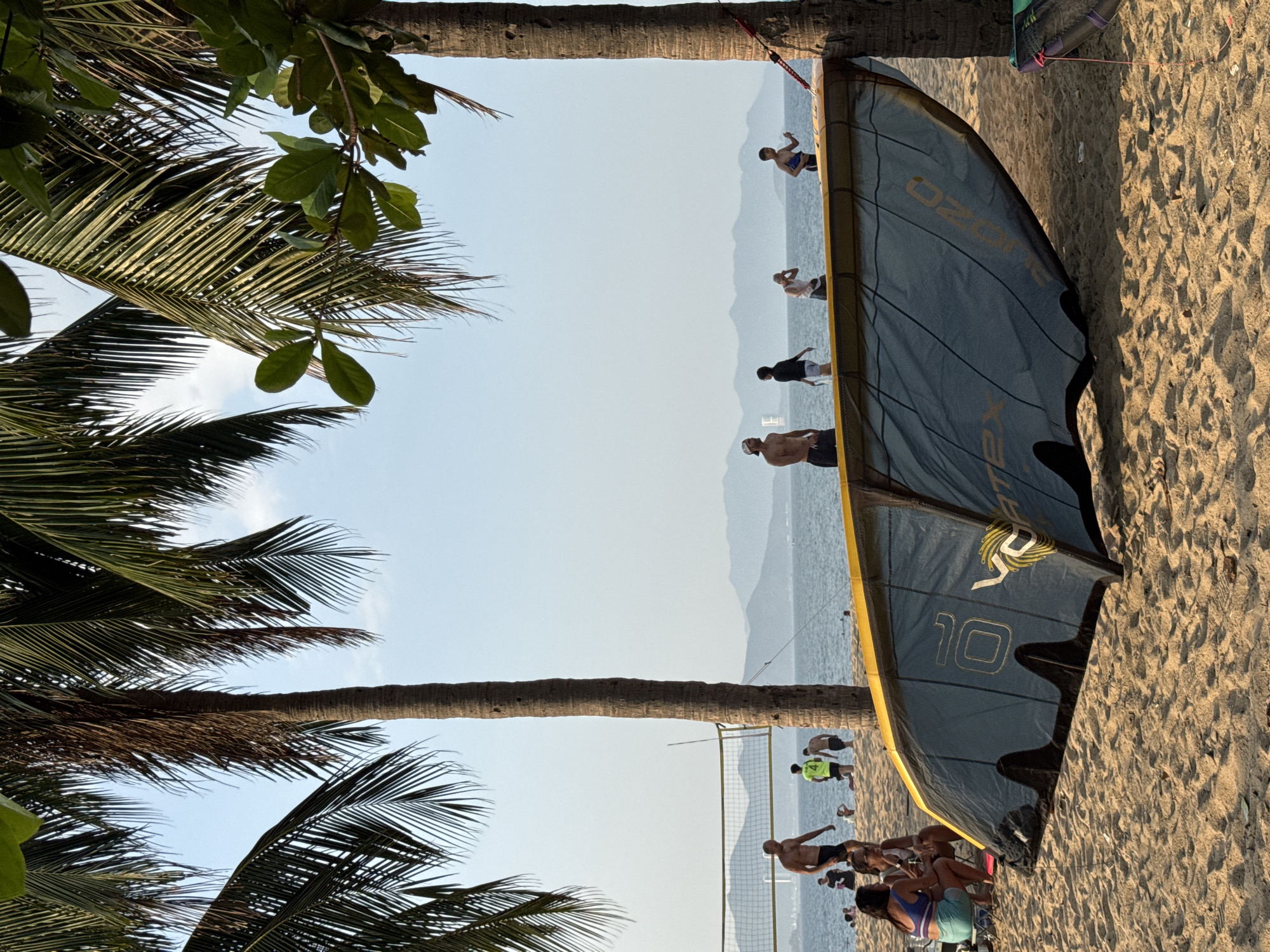 This image depicts a sunny day at a beach in Nha Trang, Vietnam. In the foreground, a large, grey and yellow kite lies on the sand, partially obscuring the view of the ocean. The kite is angled towards the right, with its brand name "OZONE" visible. Two women are seated on the sand to the left of the kite, facing away from the camera and appearing to be in conversation.
Further back on the sandy beach, several people are visible. A volleyball net is set up, and a few individuals are playing or standing around it. In the middle ground, various people are scattered across the sand, some walking, some standing, and some possibly wading in the shallow water. The ocean stretches out behind them, meeting a hazy horizon line where distant mountains are faintly visible.
Palm trees dominate the upper portion of the image, their fronds casting dappled shadows on the sand. The sky is a clear, pale blue. The overall scene suggests a recreational atmosphere on a warm day at the beach.