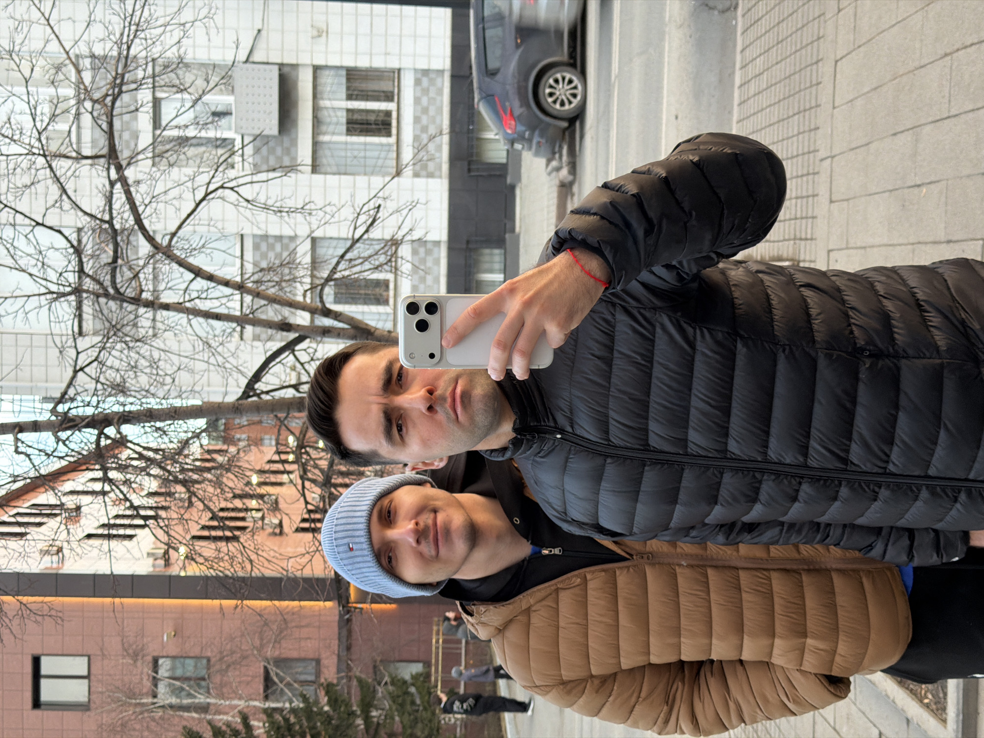 Two men in winter jackets are taking a selfie with a smartphone. The man holding the phone is wearing a black puffer jacket and has dark hair. The man in the background is wearing a light blue beanie and a tan puffer jacket. Behind them are buildings with windows and bare trees. A car is visible on the street. The scene is likely in an urban environment. Based on visual cues, the location is likely Krasnoyarsk, Russia.