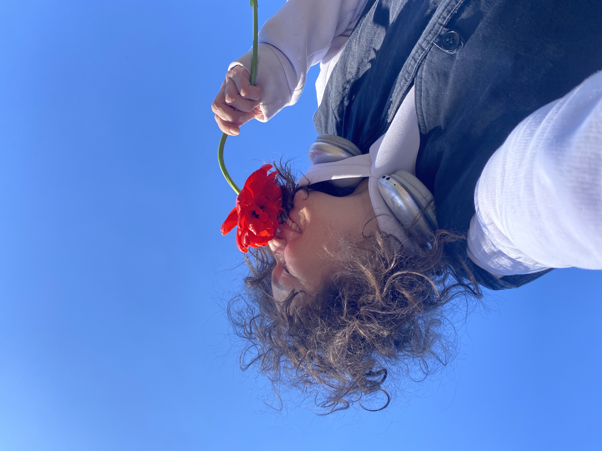 A low-angle shot depicts a person holding a red flower to their mouth. The person has curly brown hair, a white long-sleeved shirt, and a dark jacket. Headphones are draped around their neck. The background is a clear blue sky. The location is Ольховка, Russia.