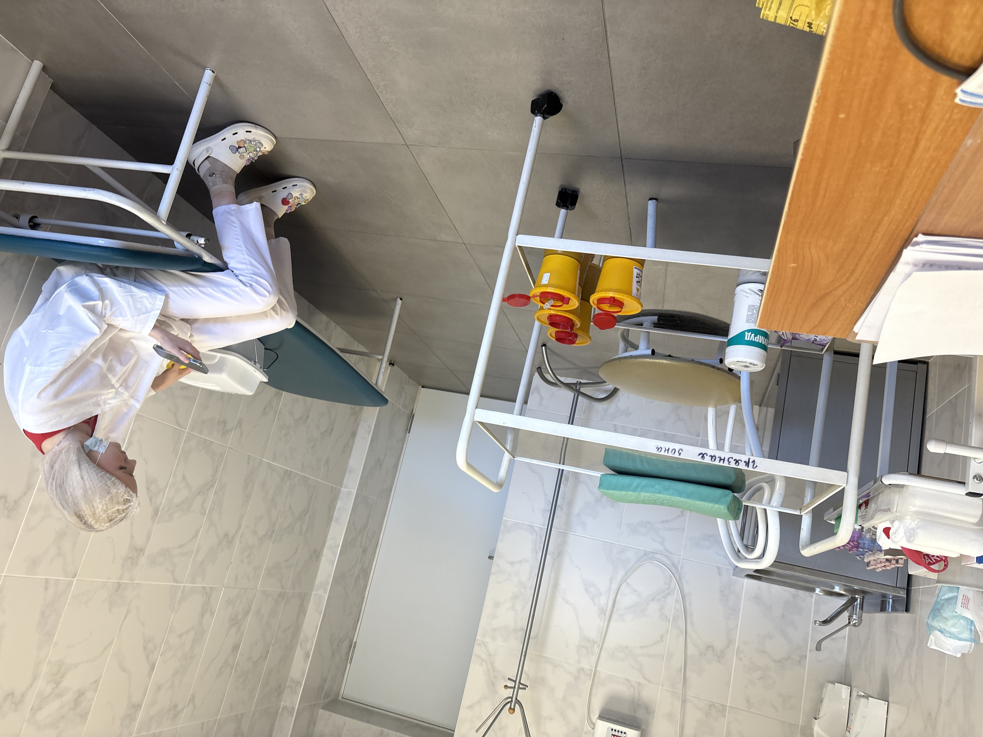 A medical examination room in Saint Petersburg, Russia. A female medical professional wearing a white lab coat, white pants, white clogs, a hairnet, and a medical mask is seated on an examination table, looking down and holding an object in her hands. The room is tiled in a marble pattern. A metal medical cart with yellow sharps containers, a metal examination table, and a sink are visible.
