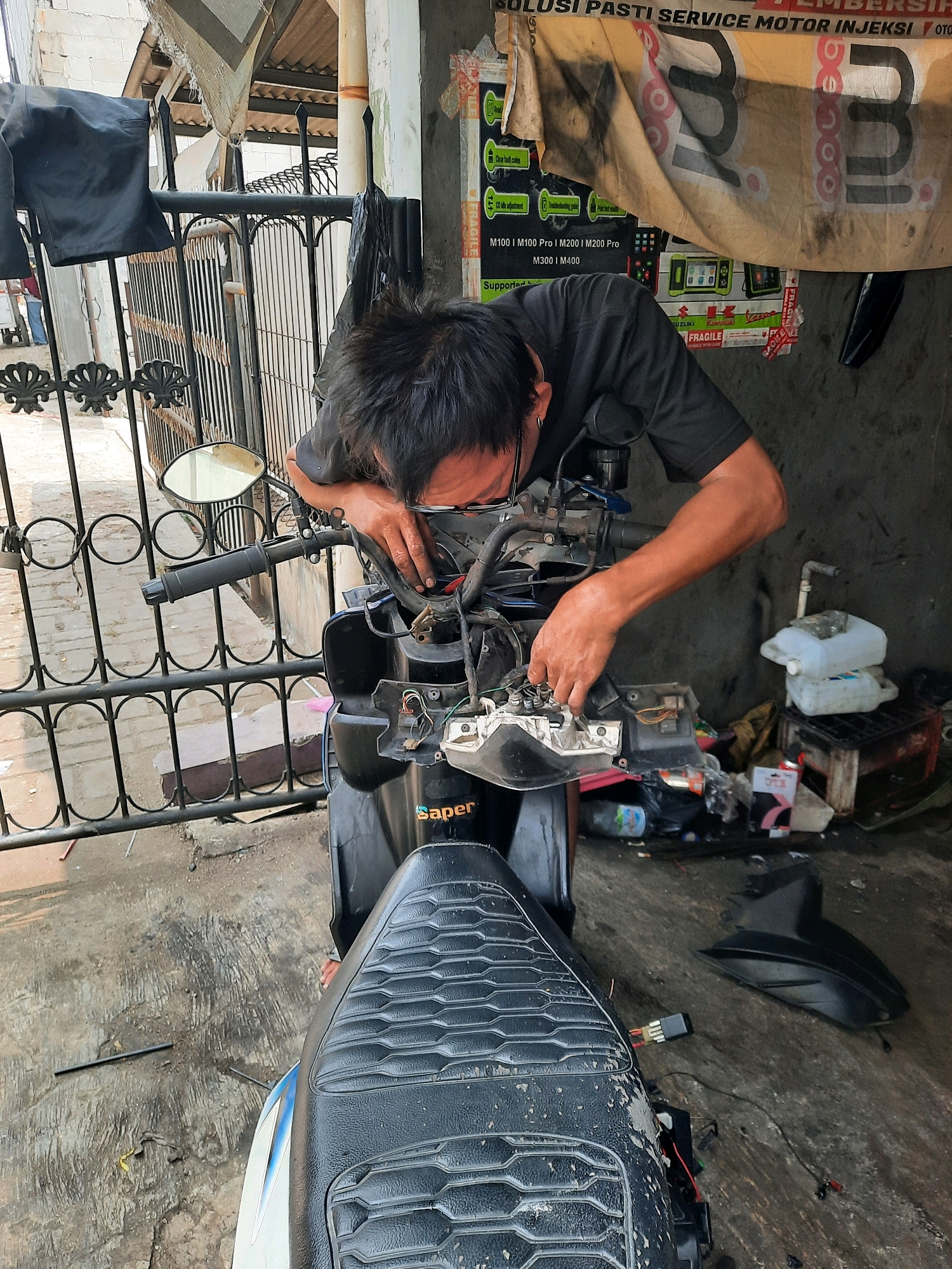 This is a medium shot of a person working on a motorcycle in an outdoor workshop. The person, wearing a black t-shirt, is leaning over the handlebars of a motorcycle and appears to be repairing or inspecting its electrical components. Wires and parts of the motorcycle's front assembly are exposed. The setting is a humble workshop with a concrete floor. Behind the motorcycle, a metal fence and a building with a corrugated metal roof are visible. Various items are scattered around the workshop, including plastic containers, bags, and what appear to be spare parts. A large banner with text in Indonesian, advertising motorcycle service, hangs on the wall behind the person. The text on the banner includes "SOLUSI PASTI SERVICE MOTOR INJEKSI OTO", "M100 I M100 Pro | M200 | M200 Pro", and "M300 I M400". Other smaller text such as "Saper", "FUZUKI", and "FRAGILE" are also visible on the banner and nearby items. The lighting suggests it is daytime. The overall scene depicts a busy, functional workspace dedicated to motorcycle repair.