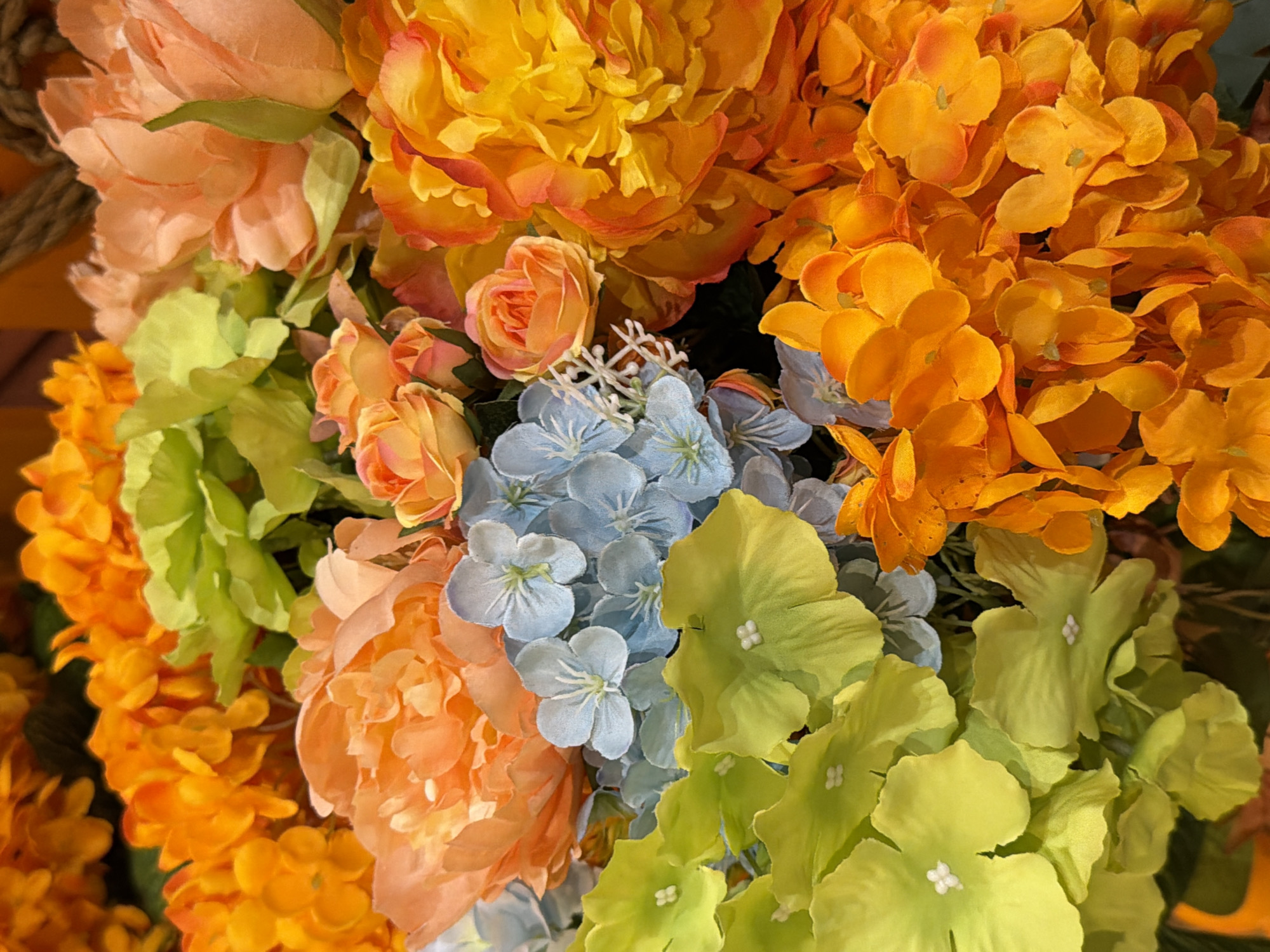 A vibrant, high-angle, close-up shot showcases a dense arrangement of artificial flowers in a bouquet. The bouquet is composed of various types and colors of flowers, including bright orange peonies and smaller orange roses, lime green hydrangeas, and delicate light blue hydrangeas. The textures of the petals appear soft and silky, with some flowers exhibiting layered petals, while others are more open and delicate. The lighting is bright and even, highlighting the rich colors and intricate details of the floral arrangement. The background is softly blurred, with hints of darker floral elements and a textured rope, suggesting the bouquet is part of a larger display or arrangement. The scene appears to be indoors, with no visible outdoor elements or specific urban context.