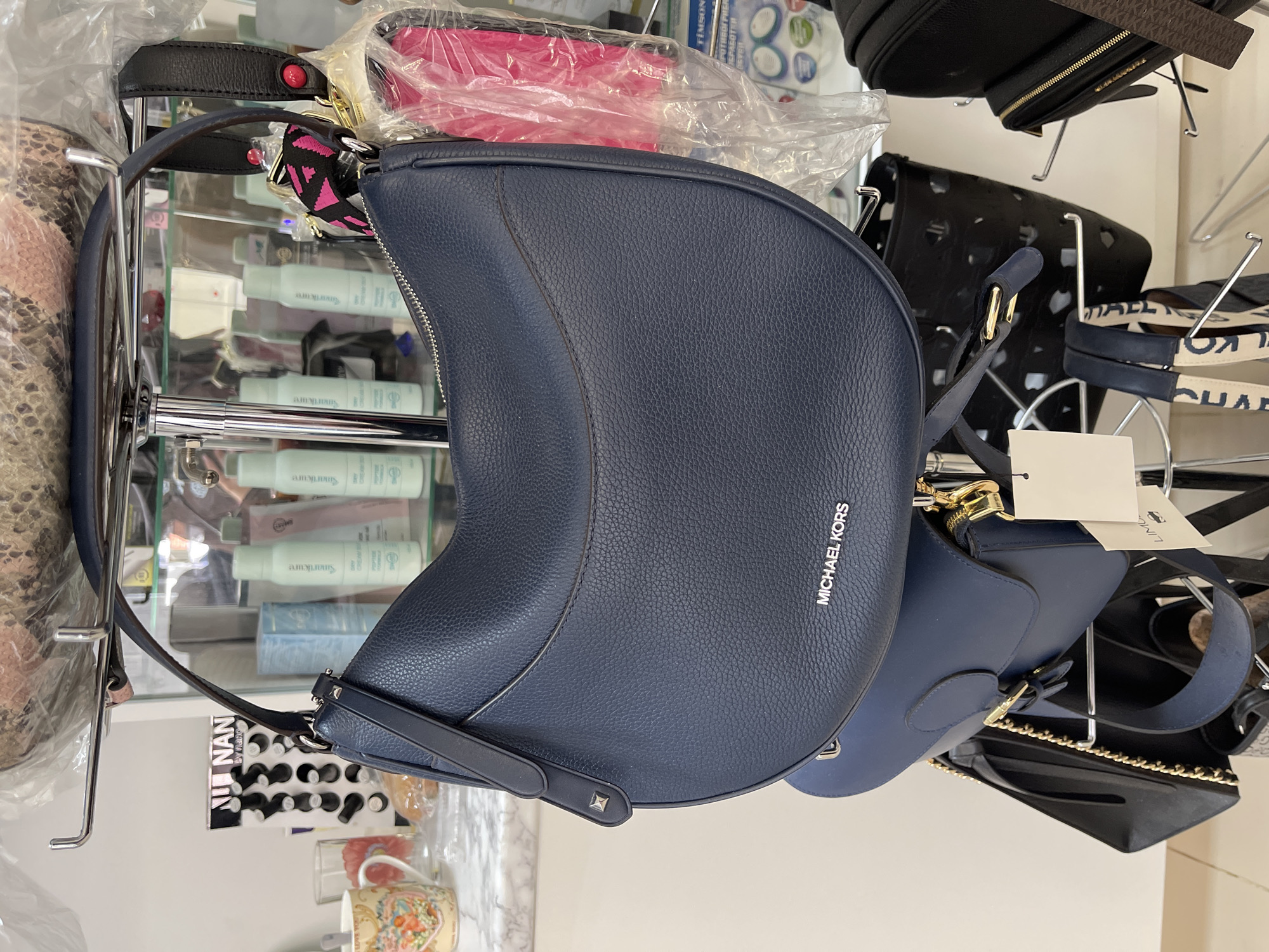 The image shows a close-up of a display rack with various handbags, likely in a retail setting. The primary focus is a navy blue Michael Kors purse hanging prominently in the center. Other purses, including a snakeskin-patterned one and a black bag with a gold chain, are visible around it. Small containers of what appear to be cosmetic products can be seen on the shelves behind the purses. A small cup and other miscellaneous items are also present in the lower part of the image.
Given that the text "TIMSON. РОТИВОГРИER ЗЕРАБОТКИ" is visible, it might be in Podolsk, Russia. The text on a small card reads "NAN by nano".