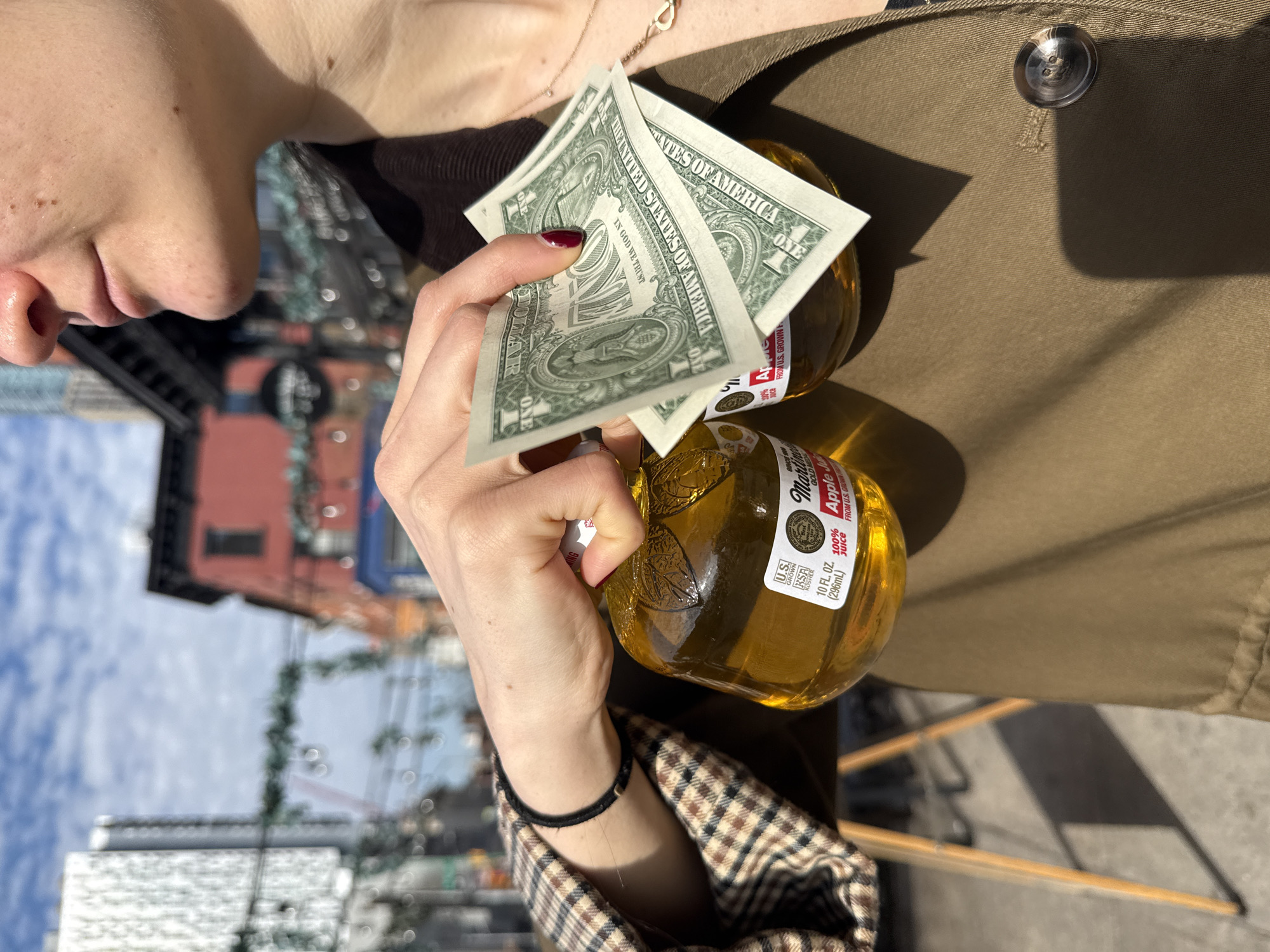 The image shows a person holding two bottles of Martinelli's Apple Juice and two one-dollar bills in front of a city street scene. The person is wearing a plaid-patterned jacket with a black cord bracelet on their wrist, as well as a brown coat. The Martinelli's bottles are glass with labels indicating they are 10 fl. oz. (296mL) and made from U.S. grown apples. The city scene in the background includes buildings with facades in shades of red, blue, and white. There is a sky with clouds visible. The location is likely New York City, United States of America, as indicated by the U.S. currency and the U.S. grown label on the juice.