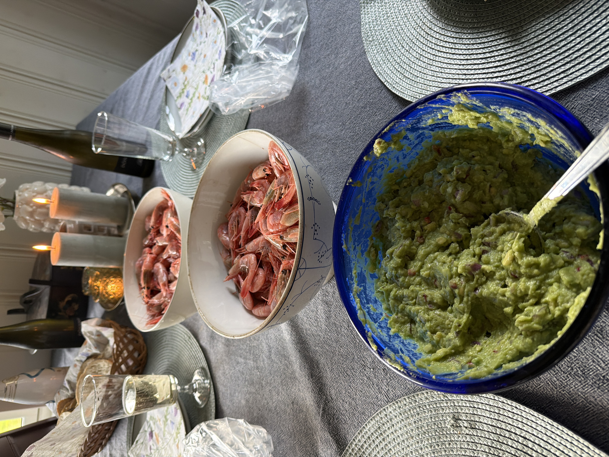 A table is set for a meal with food and drinks. In the foreground, a bright blue bowl contains guacamole, and a spoon is partially submerged in it. To the left and right of the guacamole bowl are round placemats. Above the guacamole, two white bowls are filled with shrimp. To the left of these bowls is a glass of champagne and a basket of bread. In the background, there are two lit candles, a bottle of wine, and a decorative vase with flowers. The scene is indoors, likely in a dining room. The sunlight suggests it is daytime. The mention of Kongsberg, Norway is not supported by any visual evidence in the image.
