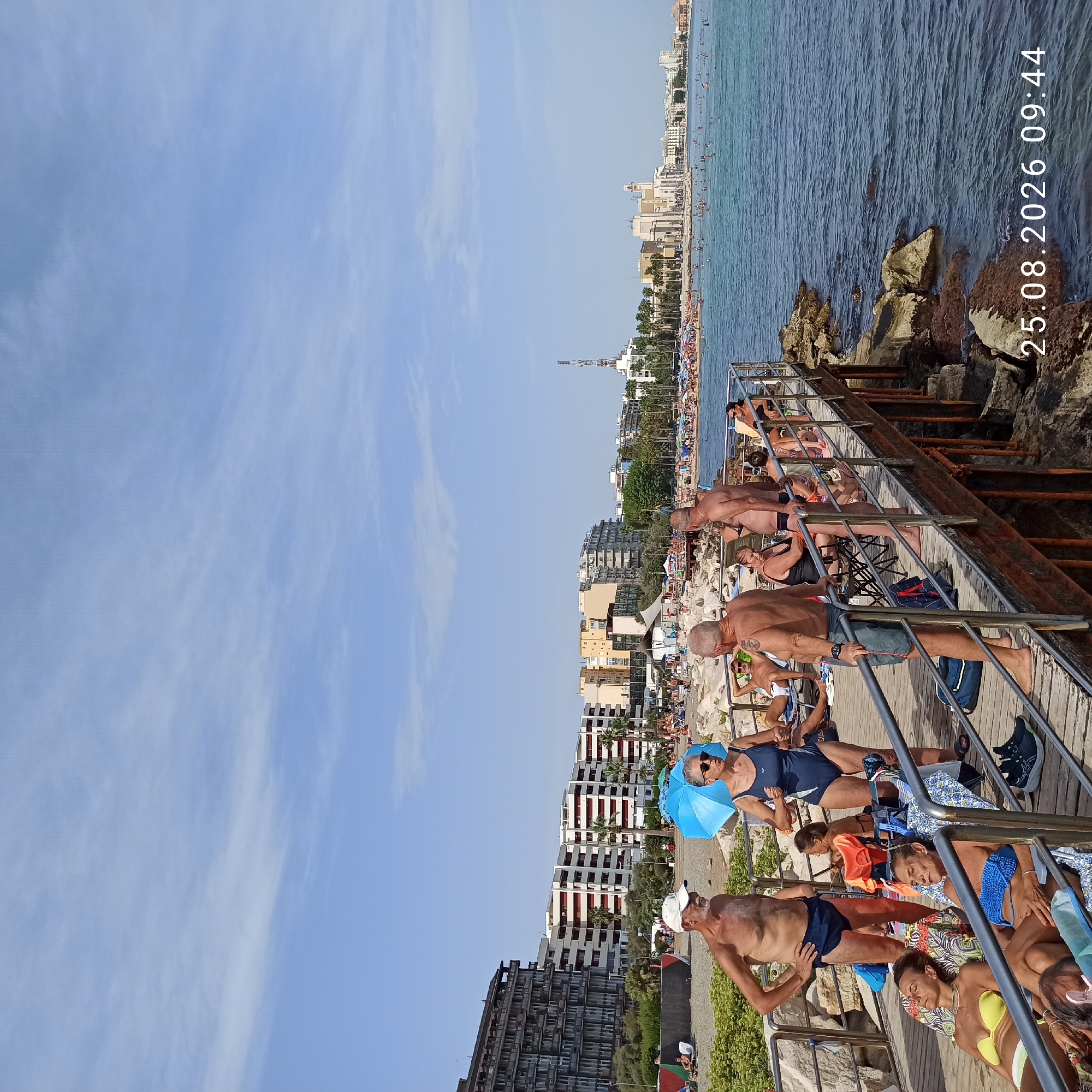 The image depicts a crowded beach and boardwalk scene on August 25, 2026, at 09:44 in Bari, Italy. In the foreground, a wooden boardwalk runs along the rocky coastline, with several individuals relaxing on lounge chairs. A man and a woman stand on the boardwalk, facing away from the camera, with other people visible in the background.
The boardwalk leads to a sandy beach filled with numerous beachgoers, sunbathers, and umbrellas. The sea is calm and a light blue, with many people swimming. In the distance, the cityscape of Bari is visible, characterized by a mix of modern and older buildings, including a prominent church-like structure. The sky is mostly clear with a few wispy clouds. The scene suggests a typical summer day at a popular tourist destination.