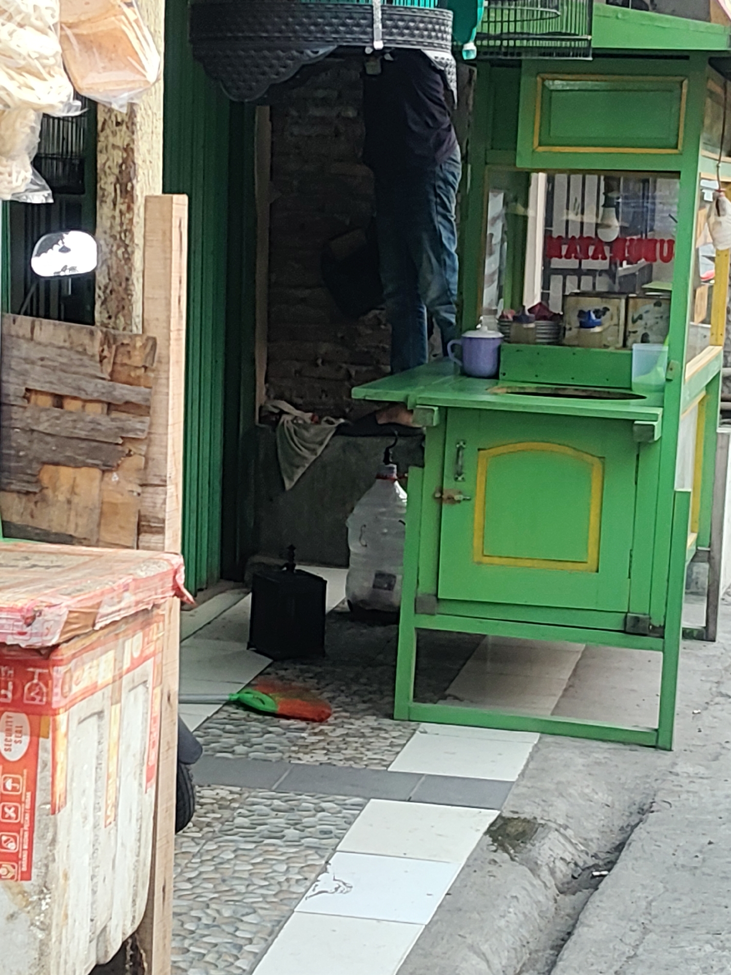 In Mekarmukti, Indonesia, an outdoor scene captures a food cart at a street-level setting. The cart is painted a vibrant green with yellow trim, featuring a display area with a glass front showcasing products. Inside the cart's display, text that appears to be "JALA JALA" is visible. A person, dressed in a dark top and blue jeans, is partially visible near the top of the cart, seemingly engaged in an activity related to the cart. On the counter of the cart, a purple mug and a clear plastic cup are present. Below the counter, a large water jug is situated on the ground, and a black bird cage sits nearby. A broom with a green handle and orange bristles lies on the ground in front of the cart. To the left of the cart, a stacked wooden structure or crates are visible, with some packaging bearing red and white markings and symbols. The ground is paved with a patterned mix of stones and square tiles. A drain runs along the edge of the paved area.