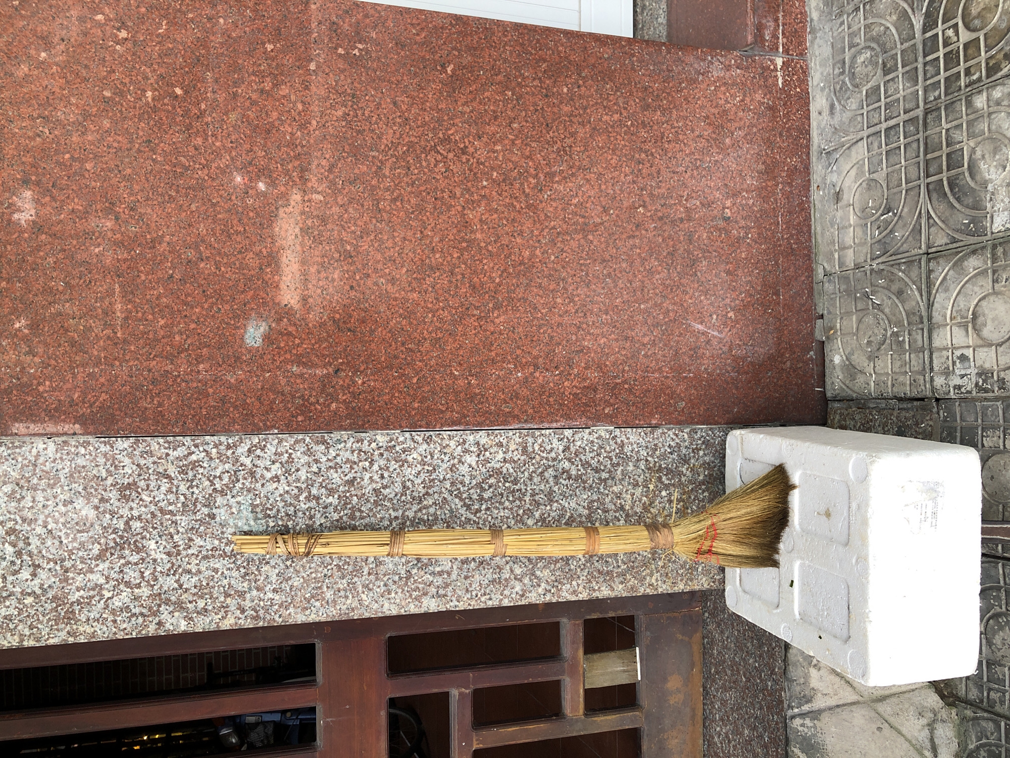 A broom made of natural fibers and bound with twine stands upright, resting on a white styrofoam block. The broom is positioned against a wall clad in speckled granite and a large panel of red granite. To the left, a wooden door with glass panes is visible. The ground is paved with patterned square tiles. The scene appears to be an outdoor location, possibly a doorway or a storefront in the city of Đà Nẵng, Vietnam.