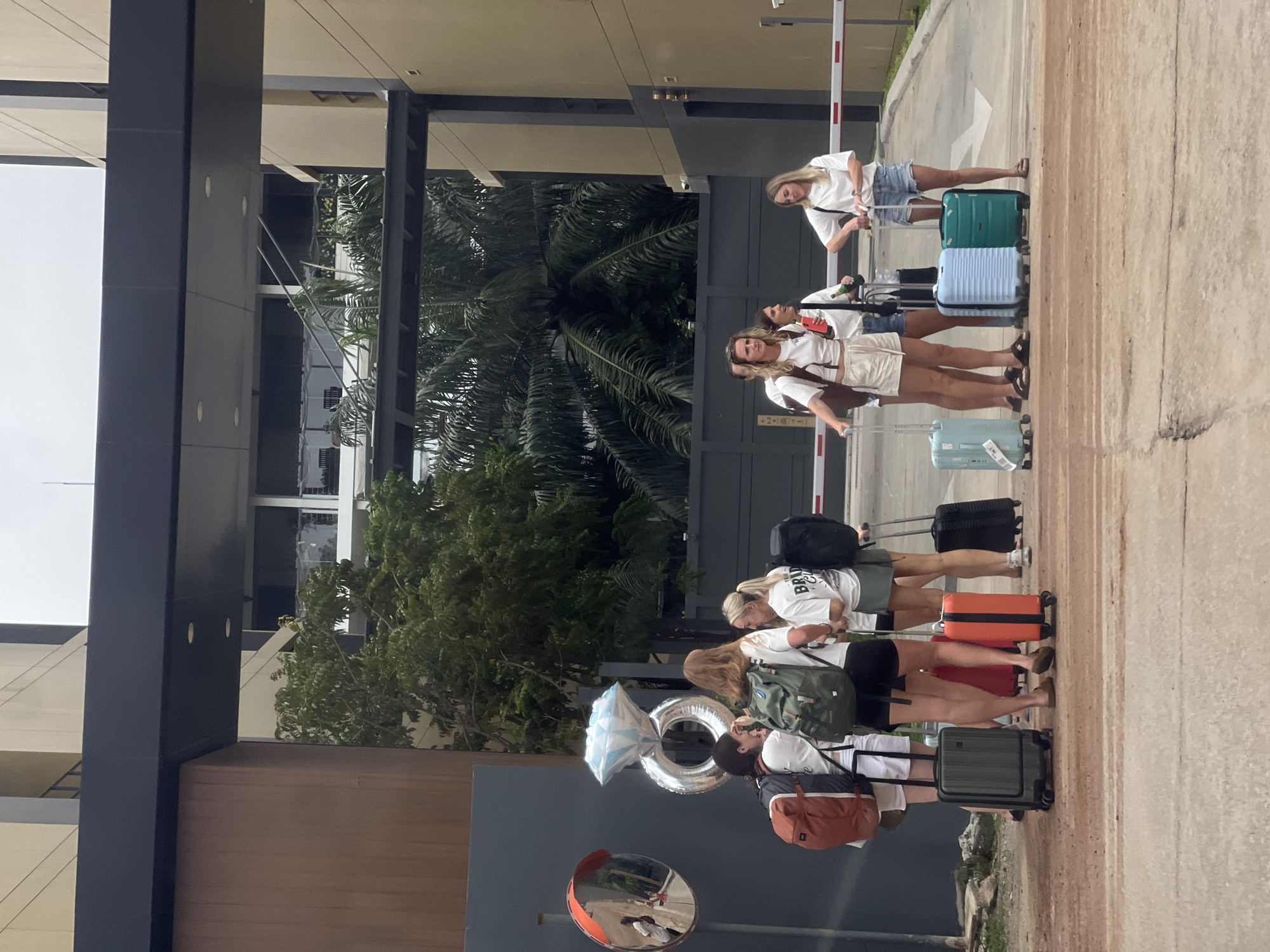 A horizontal, eye-level shot shows five women with luggage waiting next to a barrier on a sidewalk in an urban setting. The women, all wearing white t-shirts and varying shorts, are lined up with their suitcases. From left to right, the first woman has a large silver ring balloon, a rust-colored backpack, and a green suitcase. The second woman has a green backpack and a black suitcase, followed by the third woman with a black backpack and orange suitcase. The fourth and fifth women have a light blue and green suitcase, respectively. Behind the group are palm trees, and a modern building is visible on the left.