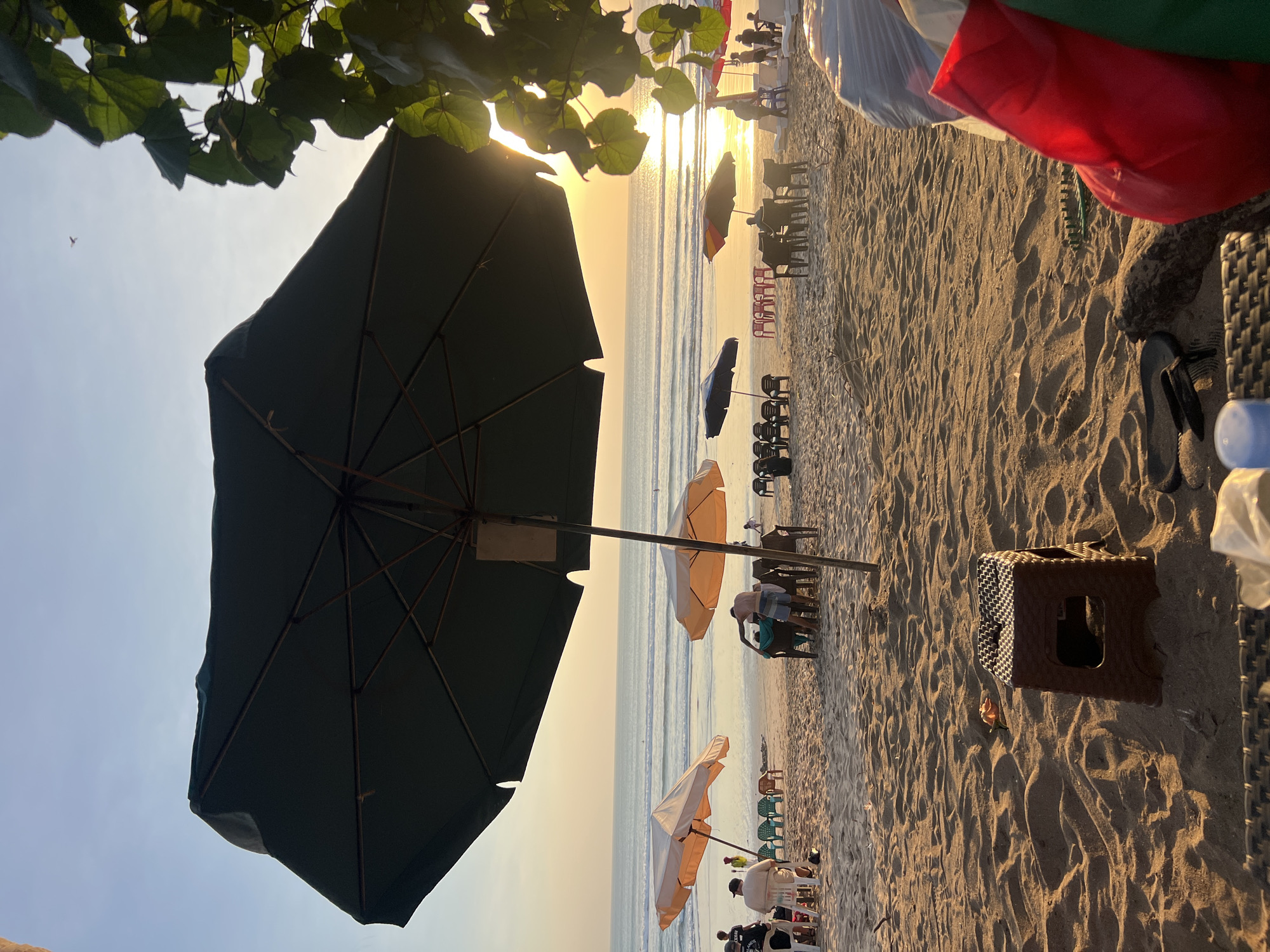 A wide-angle shot captures a beach scene in Kuta, Indonesia, at sunset. In the foreground, a large, dark green beach umbrella dominates the frame, its metal ribs visible against the pale sky. To the right, the sun's golden rays are visible through the leaves of a tree branch. The sandy beach is scattered with various beach chairs and umbrellas. Several people are seated or standing on the sand, some gathered around tables, while others appear to be walking or interacting near the water's edge. The ocean stretches to the horizon, with gentle waves rolling in. The lighting suggests the warm glow of a setting sun, casting long shadows across the sand. In the immediate foreground, a small brown plastic stool and what appears to be a pair of flip-flops are placed on the sand. A pile of items covered in white plastic and a bright red bag are also visible in the lower right corner of the frame.