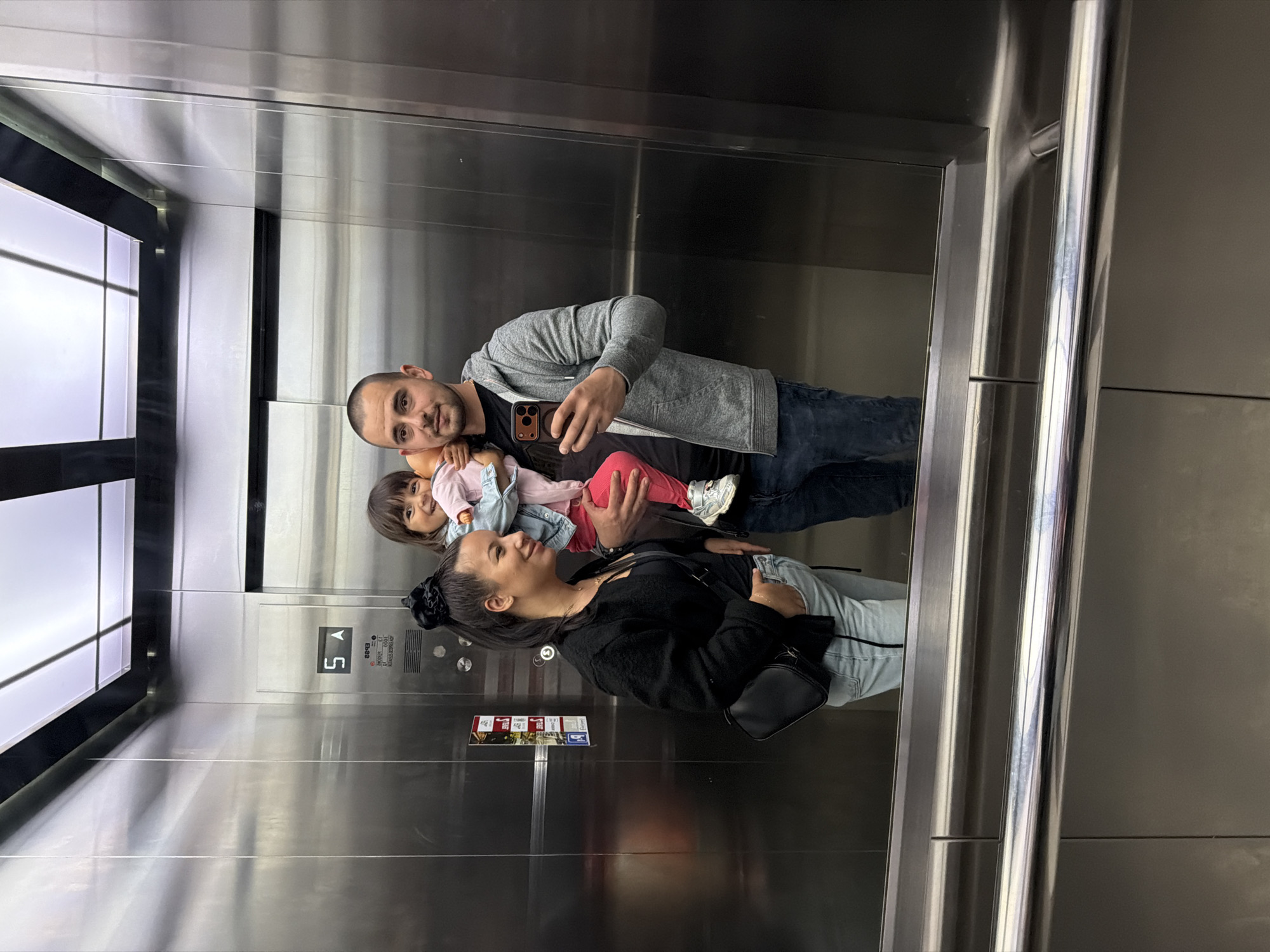 A family of three, consisting of a man, a woman, and a young child, is captured in a selfie taken inside a modern elevator in Burgas, Bulgaria. The elevator features polished, reflective stainless steel walls that mirror the subjects and the interior.
The man, positioned on the left, is taking the photo with a dark-colored smartphone, which he holds up. He is wearing a grey zip-up hoodie over a black t-shirt and dark blue jeans. He has short dark hair and a beard, looking directly at the camera with a soft smile. He holds a small child, likely a toddler, in his left arm. The child, with brown hair, is wearing a light pink top and a blue denim jacket, with visible white sneakers. The child is also looking towards the camera with a gentle smile.
To the right of the man and child, the woman stands, wearing a black long-sleeved top, light blue jeans, and a small black cross-body bag. Her dark hair is tied back with a black scrunchie. She is smiling, looking towards the man and child rather than directly at the camera.
Visible elevator features include a control panel on the wall, displaying "5" and "A," likely indicating the current floor and an operational mode. Below this, a small logo "D-20" is visible, along with an undecipherable multi-colored informational sign. A metallic handrail runs horizontally along the right wall. The bright, artificial lighting inside the elevator highlights the reflective surfaces and the cheerful expressions of the family.