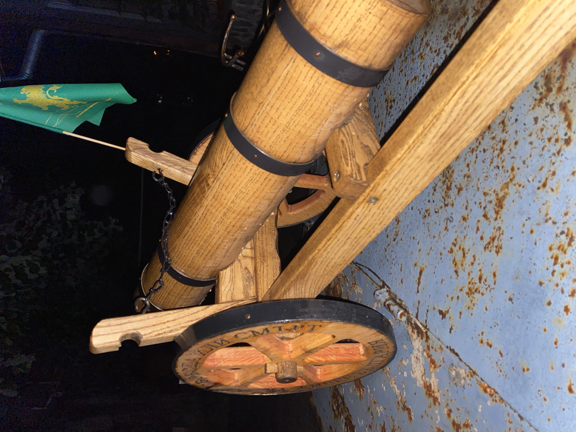 The image shows a wooden cannon model with a green flag attached to its side. The cannon is mounted on a wooden frame with wheels. The wheels have writing on them, reading "CMT-PT." The cannon is positioned next to a rusted metal wall. The location is Konstantinovo, Bulgaria.