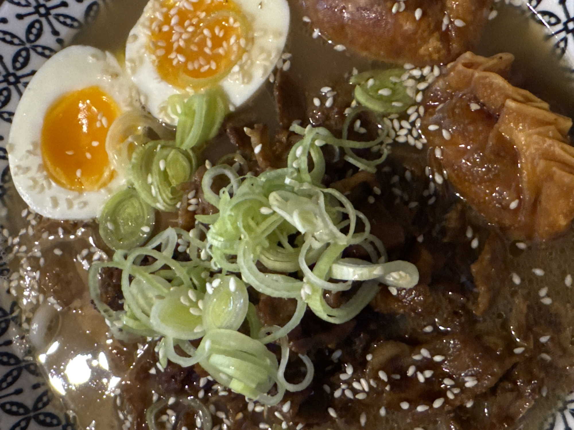 This close-up, overhead shot features a bowl of what appears to be ramen or a similar savory noodle soup. The dish is richly garnished with several key components. Two halves of a perfectly cooked soft-boiled egg, showcasing bright orange, semi-liquid yolks surrounded by solid white, are prominently displayed on the left side, both speckled with white sesame seeds.
Finely sliced green onions (scallions), curled into delicate rings, are scattered generously across the center of the dish, adding a fresh, green element. Beneath these, chunks of dark, glazed or braised meat are visible, submerged in a rich, dark brown broth. On the upper right, a piece of what resembles a crispy, fried wonton or dumpling, glistening with sauce, is partially submerged in the broth. Numerous white sesame seeds are sprinkled throughout the entire dish, enhancing its visual appeal and texture.
The food is served in a bowl with a distinct patterned rim, visible on the left and upper right sides, featuring a black and white or light blue intricate design. The image captures the dish in sharp detail, highlighting the contrasting textures and colors of the ingredients. No specific activity is depicted other than the presentation of a meal. The time of day is not discernible, and there are no visible people or text. The location context is Podgorica, Montenegro.
