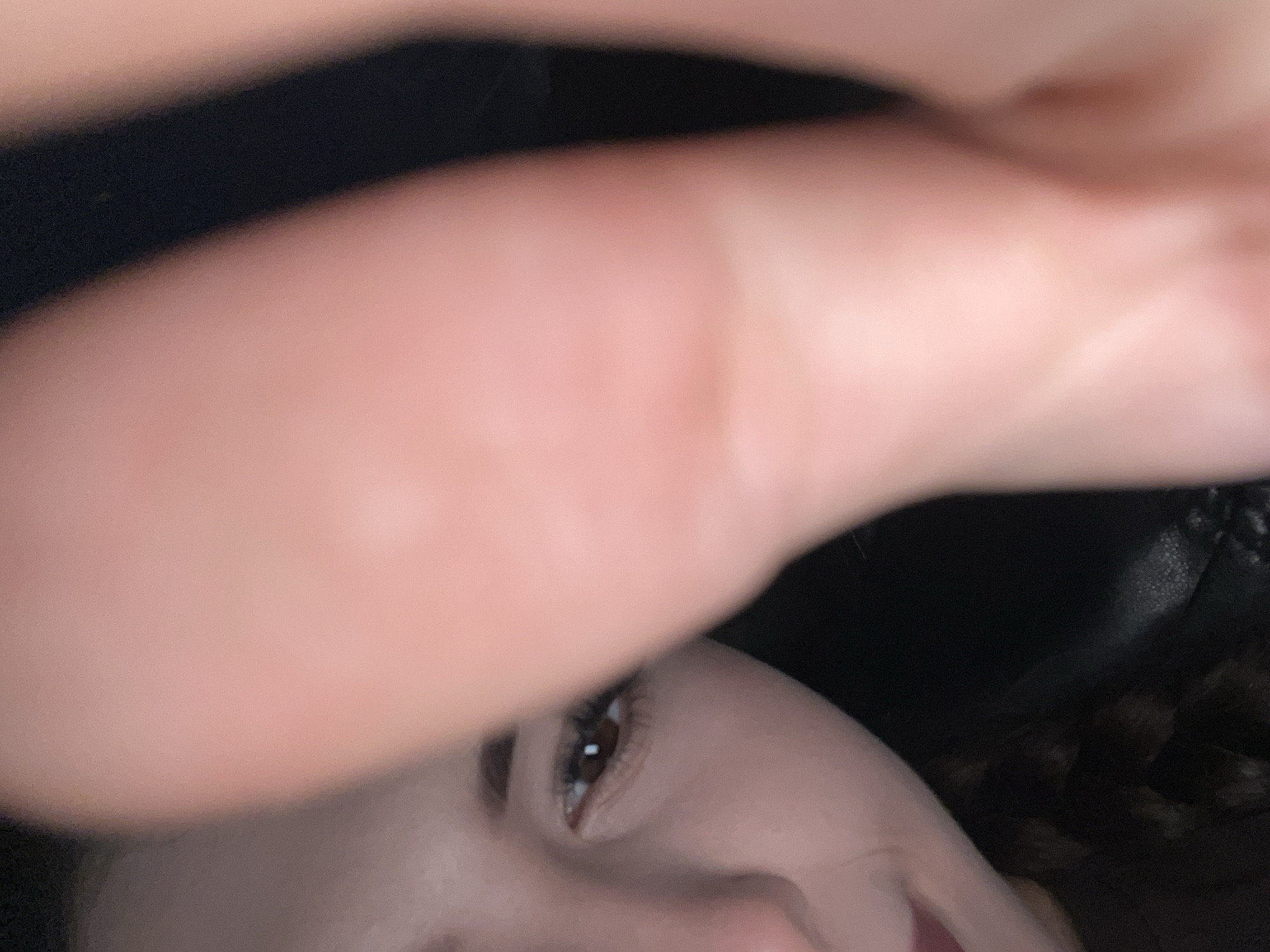 This is a close-up, low-angle, indoor, personal photograph. A portion of a young person's face is visible on the left side of the frame, focusing on their left eye and eyebrow. The person has dark hair and appears to be wearing makeup on their lips. A large, blurry, light-skinned hand or finger is prominently in the foreground, partially obscuring the view of the person's face. The background is dark and indistinct, suggesting an indoor setting with low lighting. The image appears to have been taken by the subject themselves, possibly in their residence in Пруды, Russia.