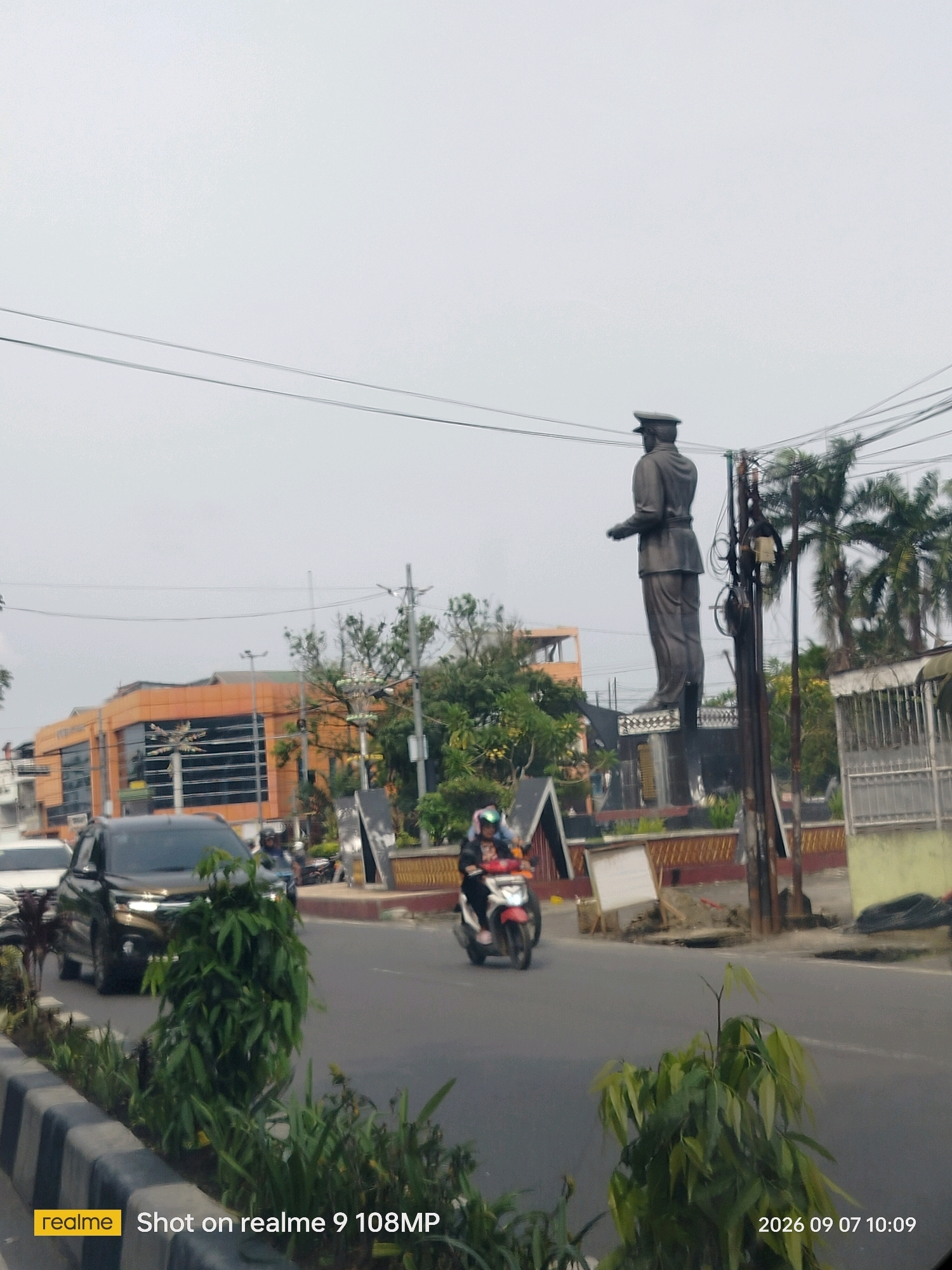 A full-shot, slightly angled, outdoor, daytime image shows a street scene in the City of Medan, Indonesia. In the foreground, a concrete median with a black and gray striped border is lined with plants. A dark-colored SUV is driving on the asphalt road to the left, while a person on a scooter is riding in the right lane. In the midground, a tall, dark statue of a man in a uniform stands prominently on a pedestal. Behind the statue, a large, orange building with dark windows is visible, along with trees and palm trees. The sky is overcast. The bottom left corner of the image displays the "realme" logo and text indicating "Shot on realme 9 108MP." The bottom right corner shows the date and time "2026 09 07 10:09."
