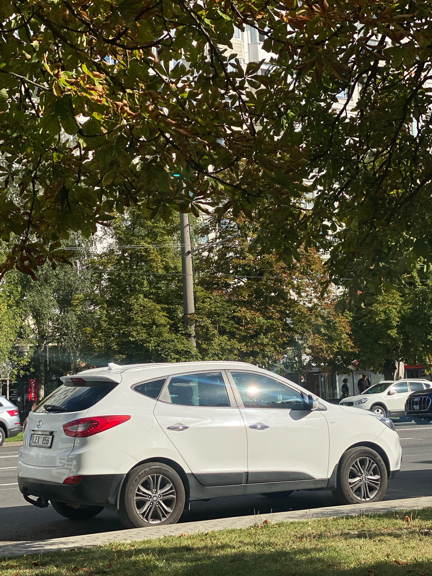 The image shows a white Hyundai SUV parked on the side of a street in Chișinău, Moldova. The car has a license plate that reads "LEX 856". The street is lined with trees, and there are buildings in the background. The sun is shining, and there are shadows cast on the street and sidewalk. There are other cars visible on the street.