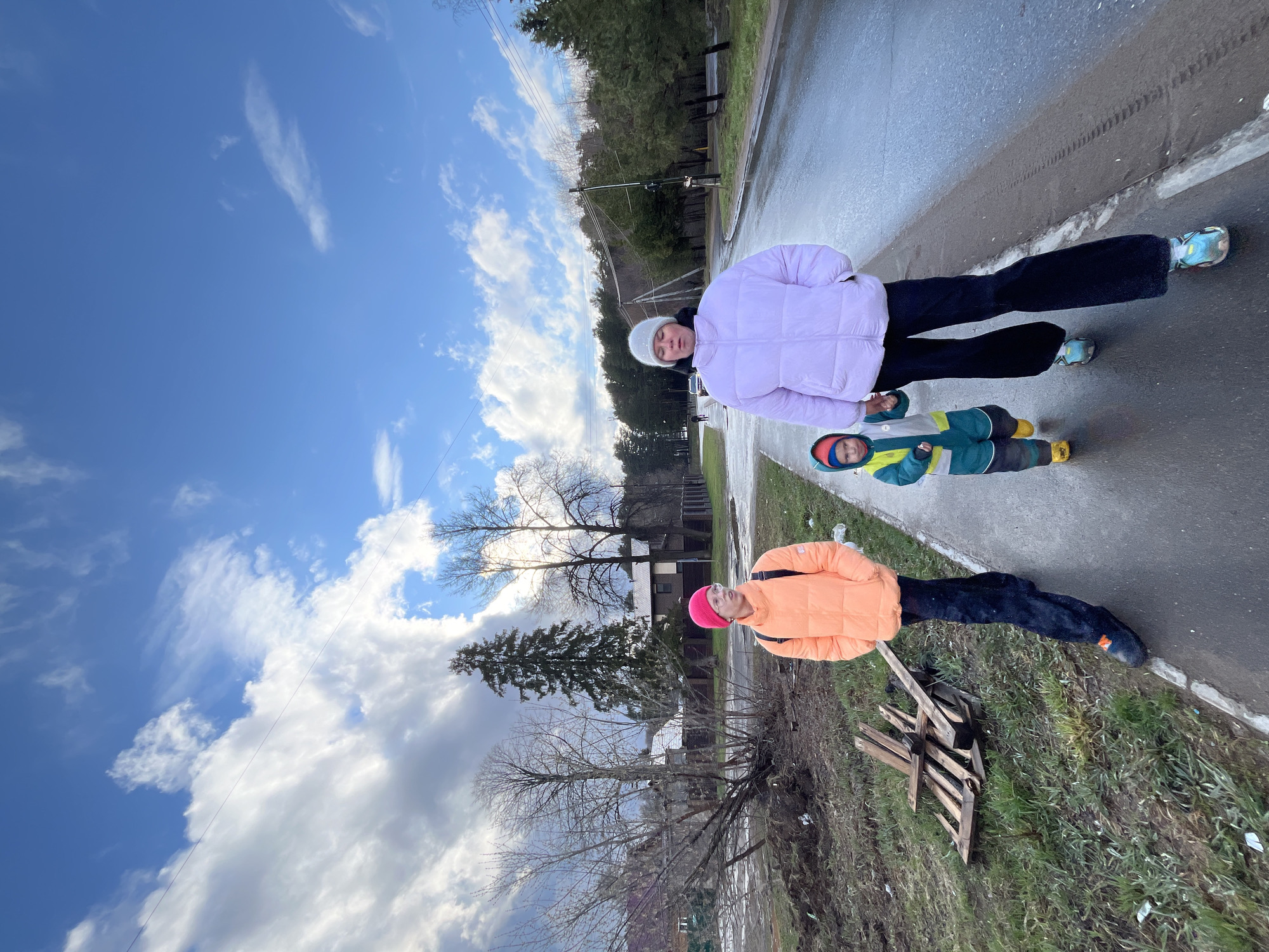A medium shot, composed at an oblique angle, depicts three individuals standing on a paved road in what appears to be the city of Chaikovsky, Russia. The upper-most person is wearing a light purple puffer jacket and a white knit hat. Below and slightly to the left is a young child in a green, blue and yellow snowsuit. Lower down and to the left is another person wearing an orange puffer jacket and a pink hat. The ground on the lower left of the image is covered with sparse vegetation and what looks like a wooden pallet. A row of trees and some houses are visible in the background. The sky above is a mix of blue sky and white and gray clouds. The paved road is wet, suggesting recent rain or melting snow.