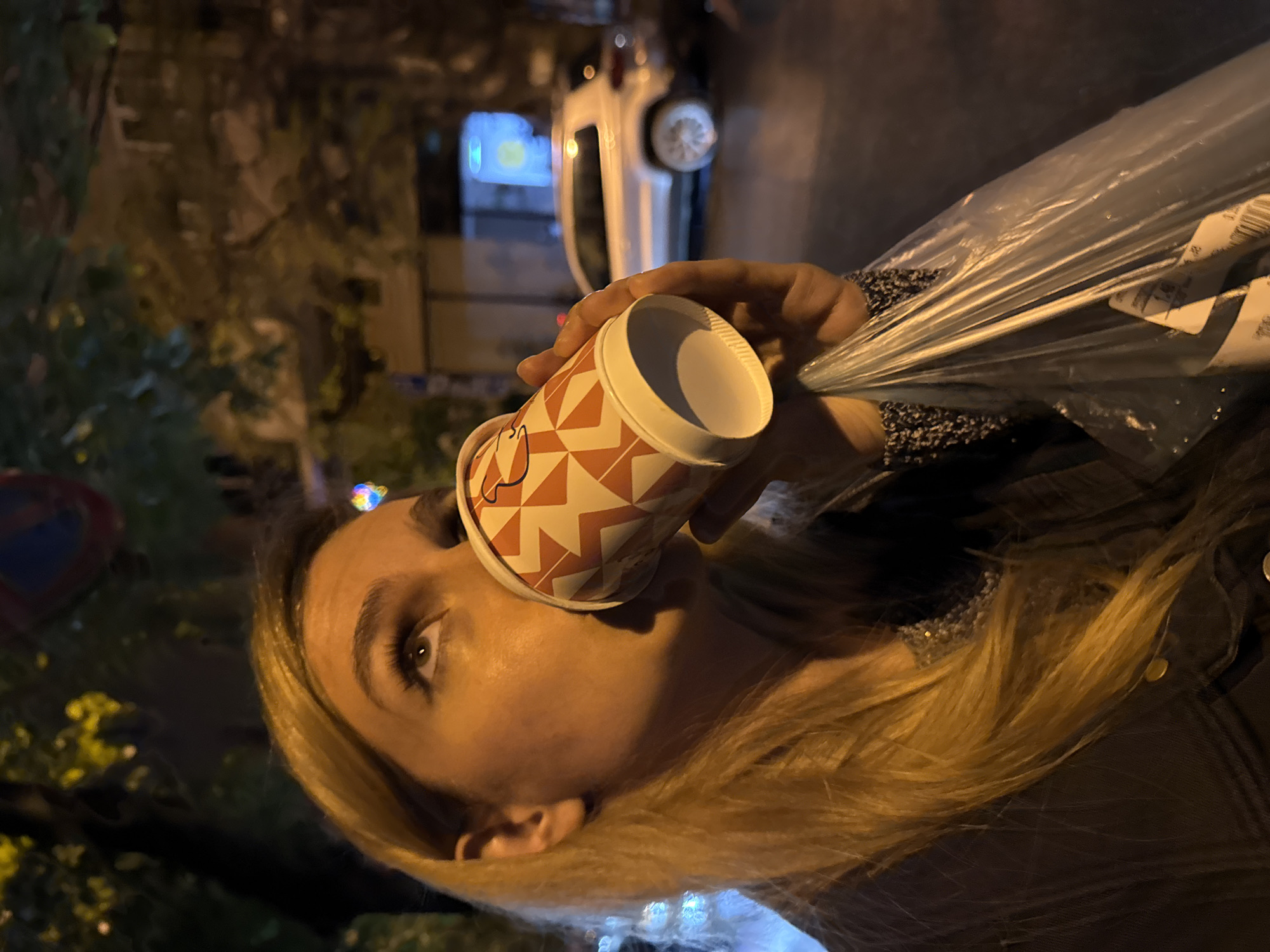 The image, oriented vertically, presents a close-up of a person outdoors at night. The main subject is a woman with long, light blonde hair, whose face is partially obscured by a paper cup she holds to her mouth. Her left eye, eyebrow, and part of her cheek and nose are visible, showing a neutral or focused expression.
The paper cup features a prominent pattern of red/orange zig-zags and triangles on a white background, topped with a white lid. A hand, likely the woman's, grips the cup. A partial word, possibly "DEL" or "GEL," is faintly visible in dark writing on the side of the cup.
Next to the woman, a transparent plastic bag is held, containing several long, thin objects, possibly art supplies like pens or pencils. Small, unreadable price tags or labels are attached to these items within the bag, with faint numbers such as "1.80" and "0.99" discernible on one tag.
The background is dark, clearly indicating nighttime. A white car, possibly a hatchback, is visible parked on a street in the mid-ground, illuminated by distant artificial lights. Green foliage from trees or bushes fills parts of the background, dimly lit. A circular sign, partially red and blue, is glimpsed on the far left. The scene suggests an urban or suburban street at night, specifically in Tbilisi, Georgia.
