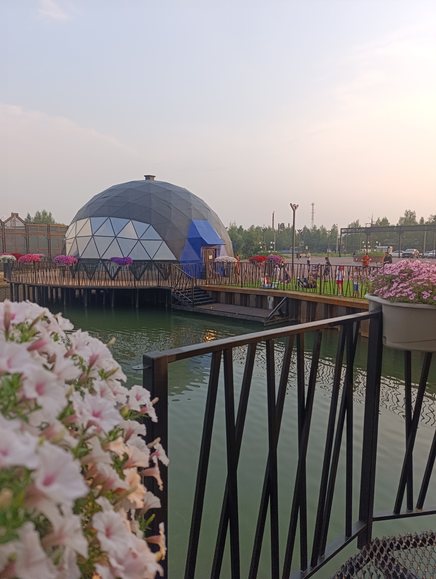 The image depicts a modern geodesic dome structure situated over water, likely a restaurant or event space. The dome has a dark grey exterior with diamond-shaped glass panels and a bright blue section that appears to be an entrance. It is surrounded by a wooden deck with black railings, adorned with planters filled with pink and purple flowers. In the foreground, a cluster of white and pink petunias blurs the edge of the frame, adding a soft, natural touch.
Beyond the dome and deck, a body of calm green water reflects the overcast sky. People are visible on the grassy area behind the deck, some standing and conversing, others seated, suggesting a relaxed social gathering or outing. Further in the background, trees and industrial structures indicate an urban or semi-urban setting. The lighting suggests it is late afternoon or early evening, with a soft, diffused light that is characteristic of an overcast day or twilight. The overall mood is tranquil and pleasant, with the natural elements of flowers and water contrasting with the modern architecture. No visible text is present in the image. The location context of Noyabrsk, Russia, suggests this might be a unique recreational spot in a northern climate.