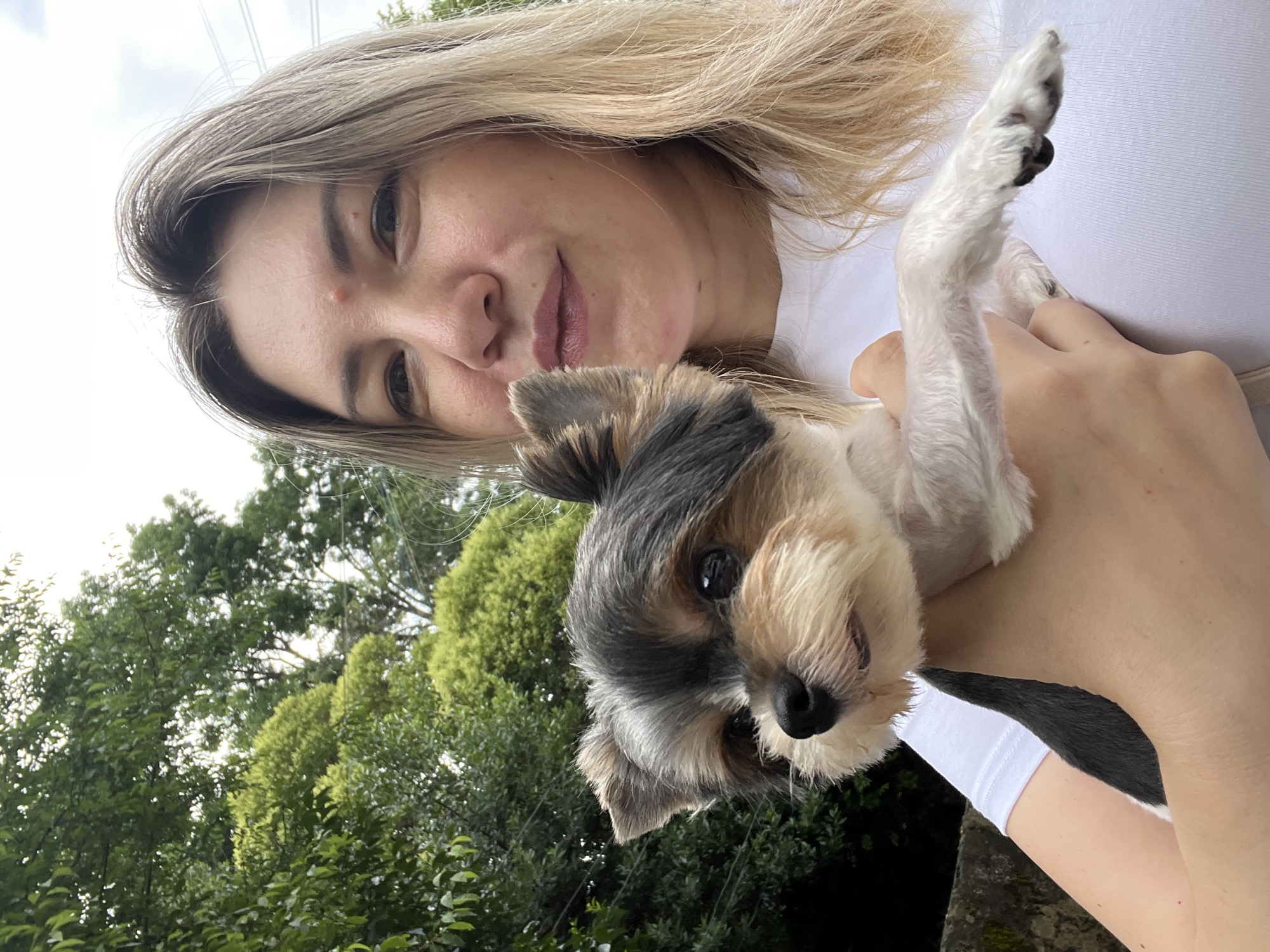 A young woman with blonde hair and a white t-shirt is holding a small Yorkshire Terrier. The dog is dark grey, black, tan, and white. The woman is taking a selfie outdoors, with lush green trees and a grey sky in the background. A stone wall is visible to the left of the woman.