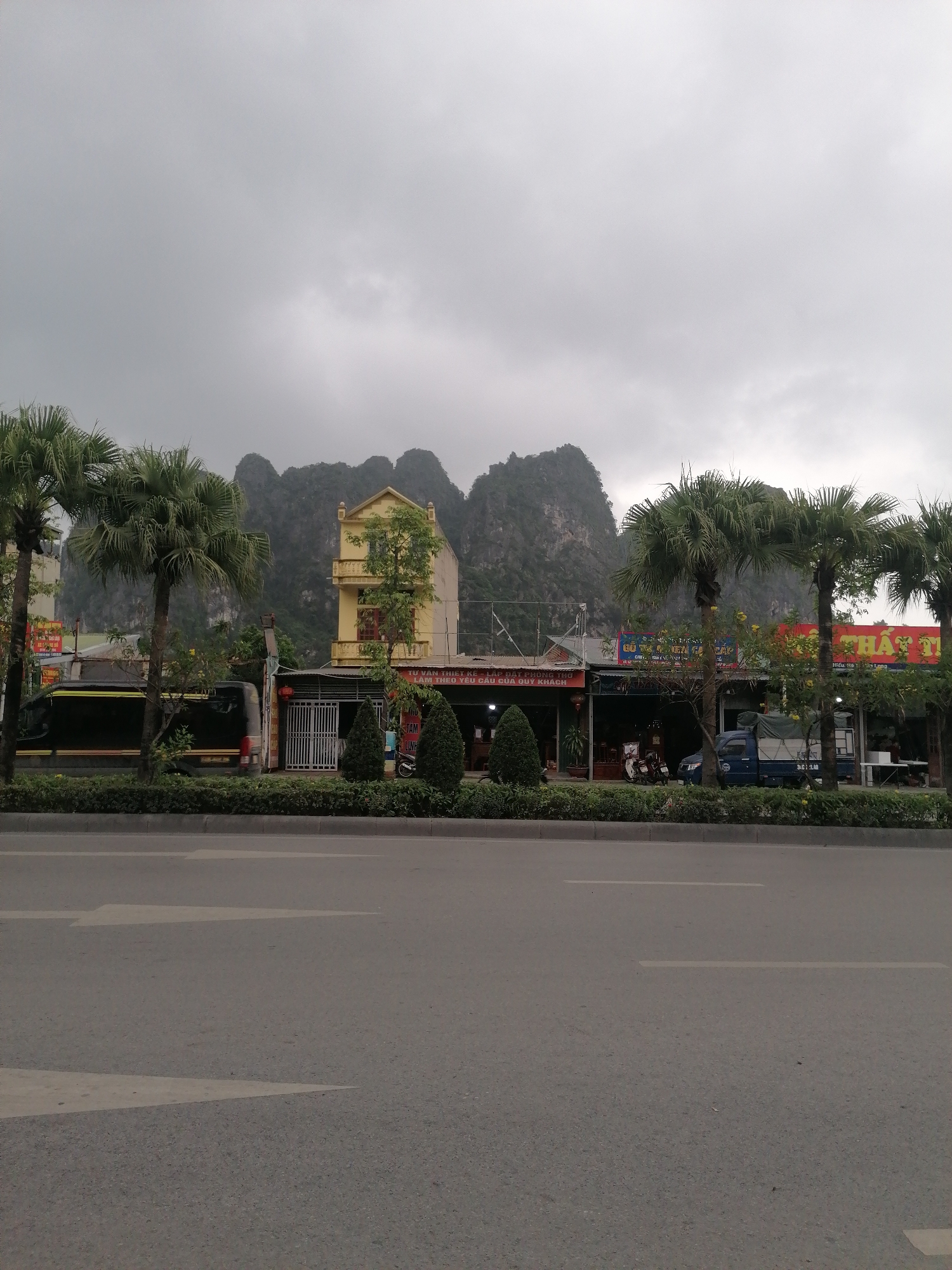 The image shows a street scene in Ha Tu Ward, Vietnam. A wide asphalt road with arrow markings dominates the foreground. Beyond the road is a low hedge, followed by a row of buildings and shops partially obscured by palm trees. One building is painted yellow and stands out against the backdrop of karst mountains, which are shrouded in a grey, overcast sky. There are parked vehicles along the street, including a black van and a blue truck. Shop signs with Vietnamese text are visible above the storefronts.