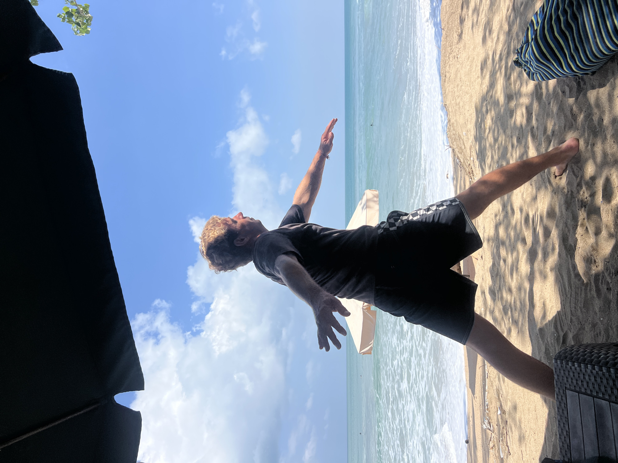 A man with blonde, curly hair is performing a yoga pose on a sandy beach in Kuta, Indonesia. He is wearing a dark t-shirt and checkered black shorts. His arms are outstretched, with his left arm extended forward and his right arm extended to the side. The ocean, with gentle waves, is visible behind him, as is a pale beige beach umbrella. The sky is bright blue with scattered white clouds. The foreground of the image is shaded by a dark umbrella, and a textured dark surface, possibly furniture, is visible on the left. A striped bag or mat lies on the sand to the right.