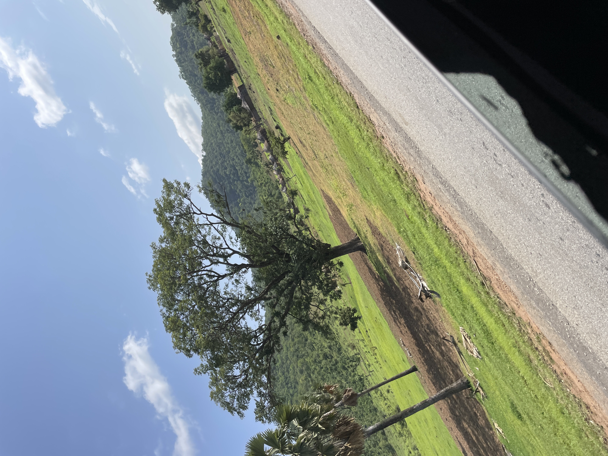 The image shows a rural landscape with a road in the foreground. The road is paved and slopes upwards to the right. On the left side of the road, there is a grassy field with scattered trees and some houses in the distance. The field is green and appears to be a pasture or farmland. In the background, there are hills covered in lush vegetation. The sky is blue with some white clouds. The overall scene depicts a peaceful, rural setting in the city of Serti, Nigeria.