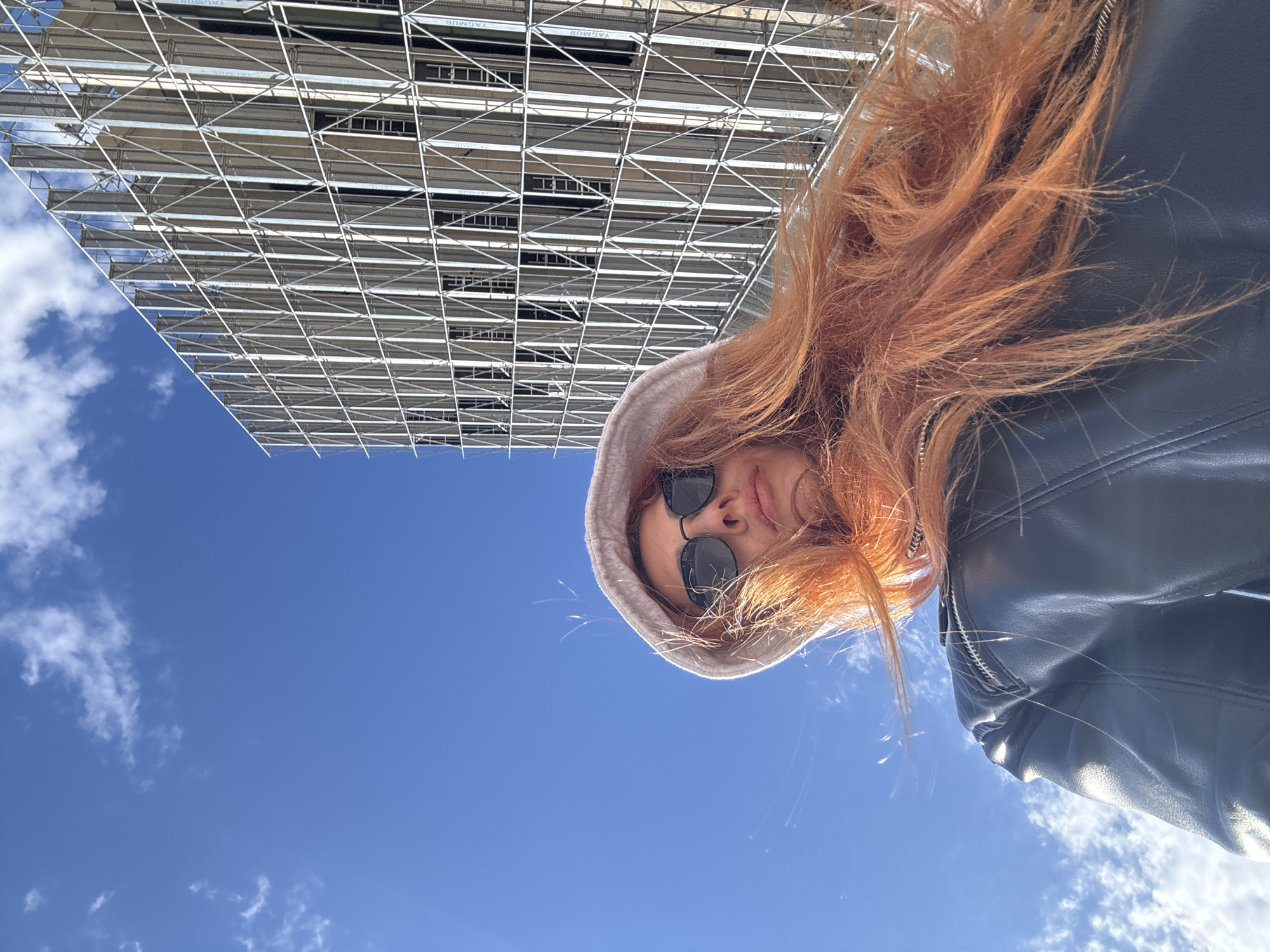 A low-angle shot shows a building, a person, and the sky. The building, constructed from metal beams, is in the upper left of the image. In the upper right, a person with reddish-orange hair wears a light-colored hooded sweatshirt, sunglasses, and a black leather jacket. The blue sky with white clouds is visible in the lower portion of the frame. The location is Budva, Montenegro.