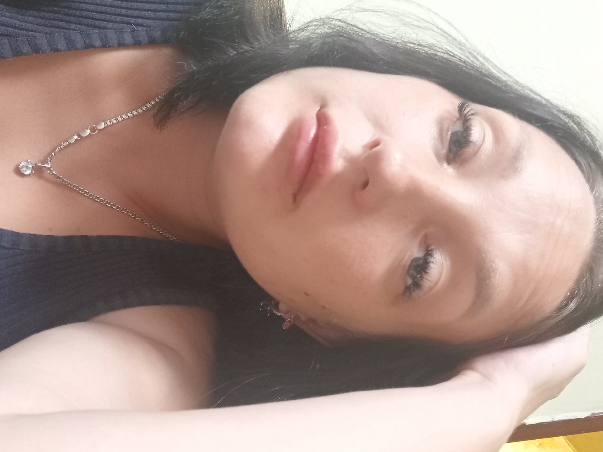 A close-up selfie shows a young woman with dark hair and light skin. She is wearing a dark blue tank top and a silver necklace with a pendant. She has her right hand on her head, and she is looking towards the camera with a neutral expression. The background is a plain light-colored wall. The image appears to have been taken indoors.
