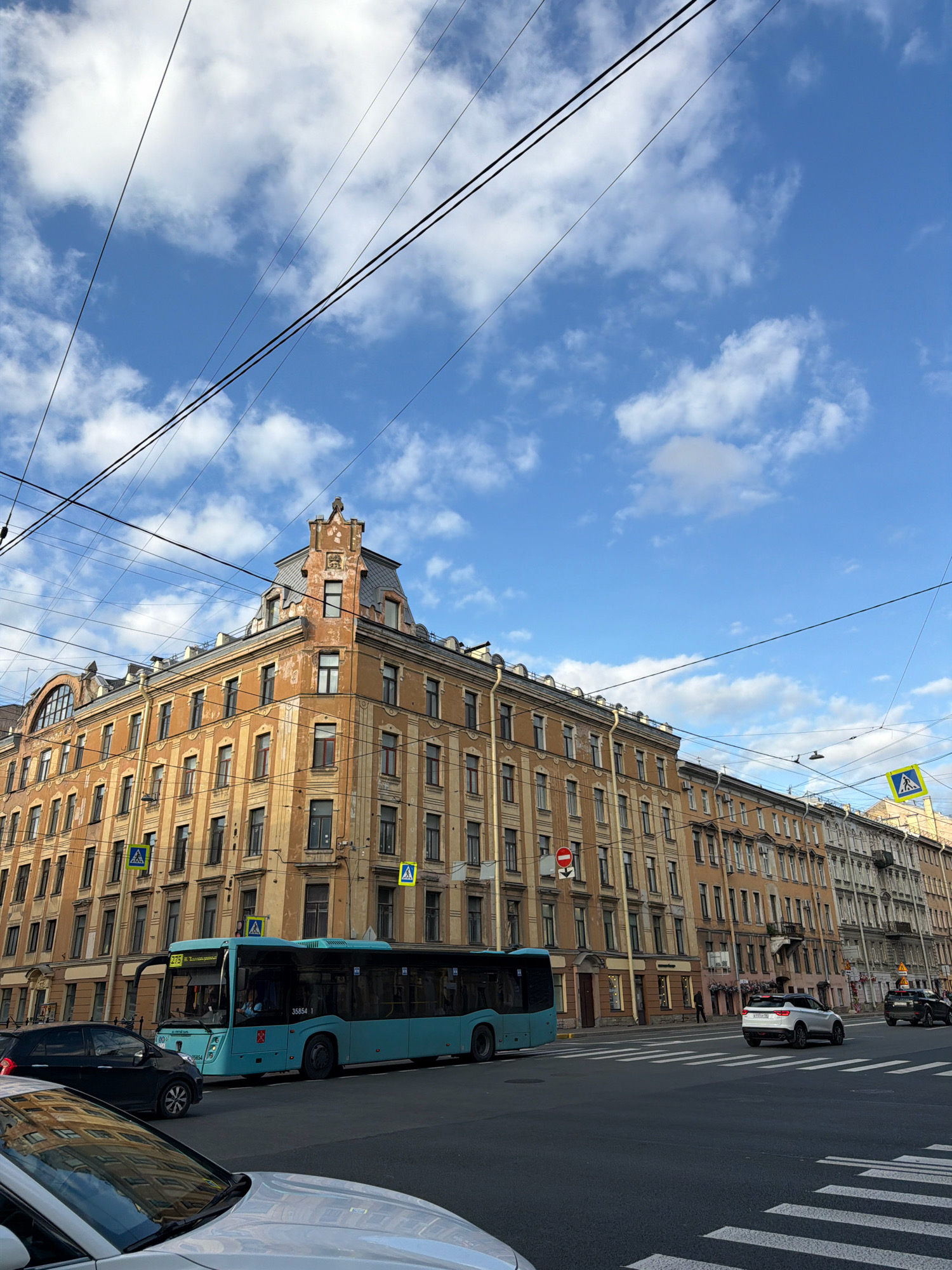 A long, multi-story, tan and yellow apartment building with a complex mansard roof and ornate architectural details dominates the scene. The building occupies the majority of the frame, stretching along a city street. In front of the building, a light blue bus travels down the street. Several cars are visible, including a white sedan in the foreground and a white SUV further down the street. A black SUV is parked on the right side of the street. A pedestrian crossing is painted on the asphalt in the foreground. Overhead, a network of utility wires crisscrosses the bright blue sky, which is dotted with fluffy white clouds. Yellow pedestrian crossing signs are visible at intersections. The building appears to be in a historic district of Saint Petersburg, Russia.