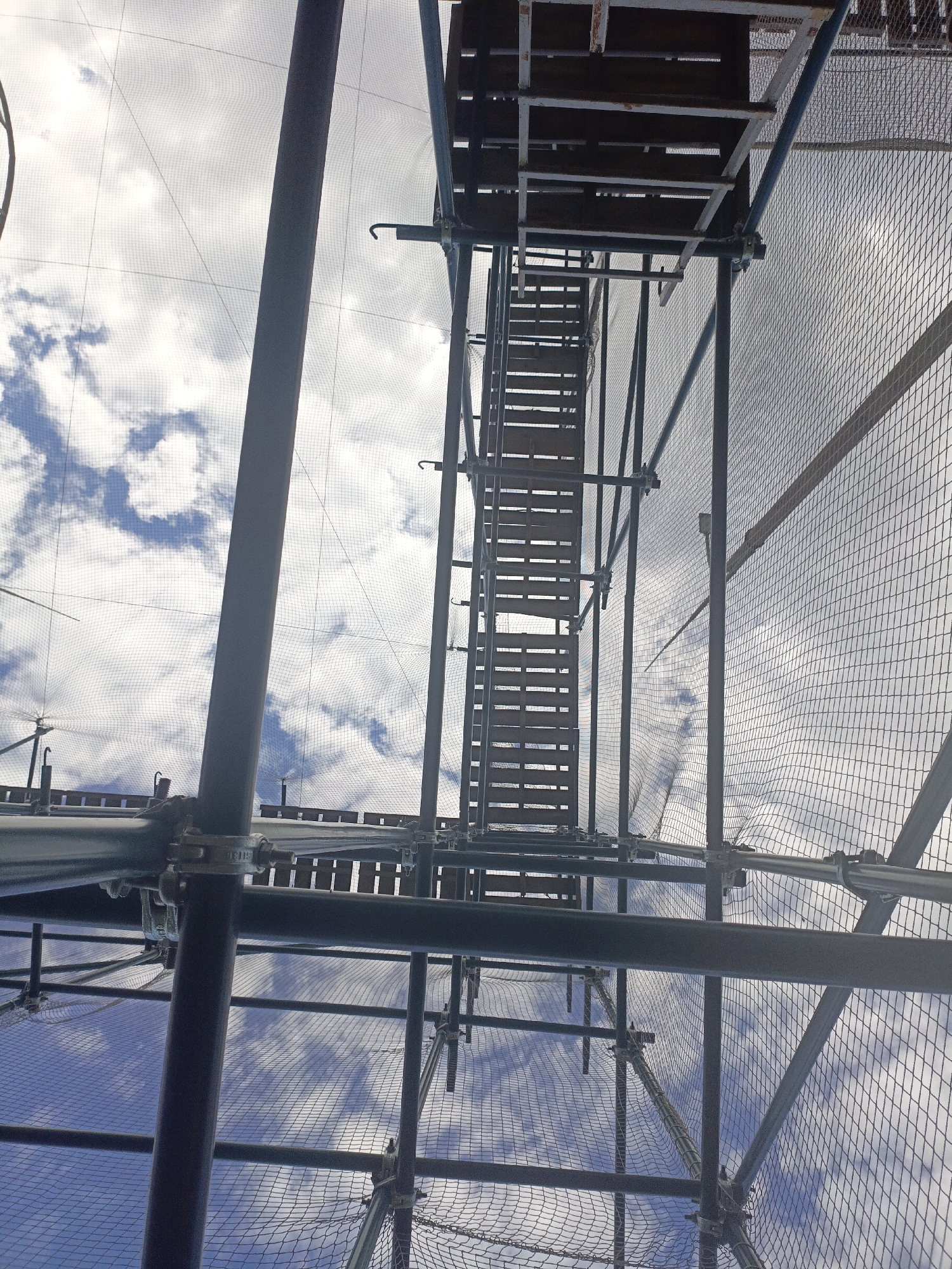 The image shows a view from below, looking up at a construction scaffolding structure against a sky with clouds. The scaffolding is made of metal poles and beams, with wooden planks forming a staircase or walkway. A fine mesh netting is stretched across the structure, likely for safety. The scene appears to be outdoors, with the bright sky and clouds suggesting daylight. There is no indication of the specific location in Ufa, Russia, nor are there any people or actions depicted. The perspective is from ground level or a lower platform, emphasizing the height and complexity of the scaffolding. The overall impression is one of ongoing construction or maintenance work.