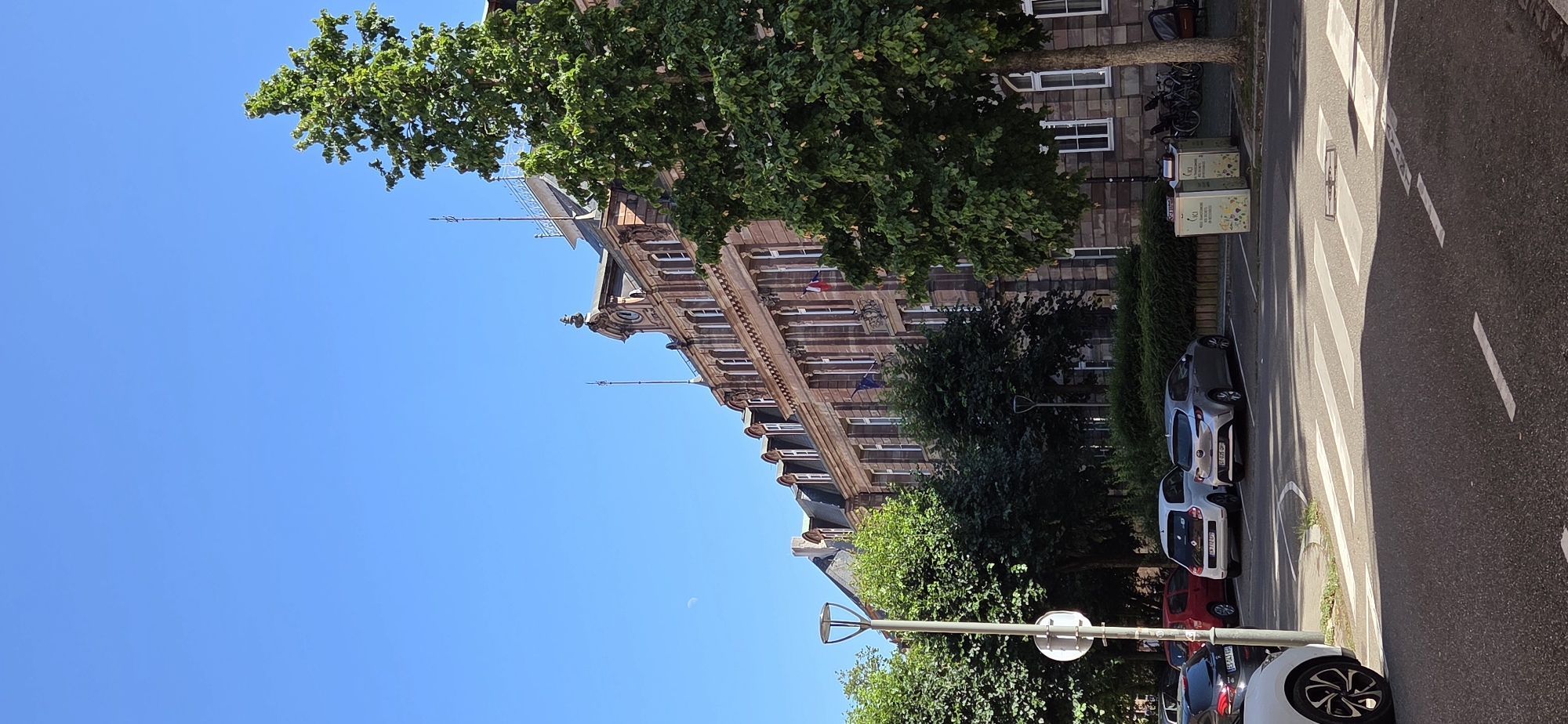 The image shows a street scene in Strasbourg, France, with a large, ornate building on the right. The building, likely a public institution or a historical monument, features red stone construction, numerous windows, and decorative architectural elements. A large green tree stands in front of the building, partially obscuring it. In the foreground, a street with a crosswalk is visible, with several cars parked on the side. The sky is clear and blue, with a crescent moon faintly visible. The scene suggests a sunny day in a city with a rich architectural heritage.