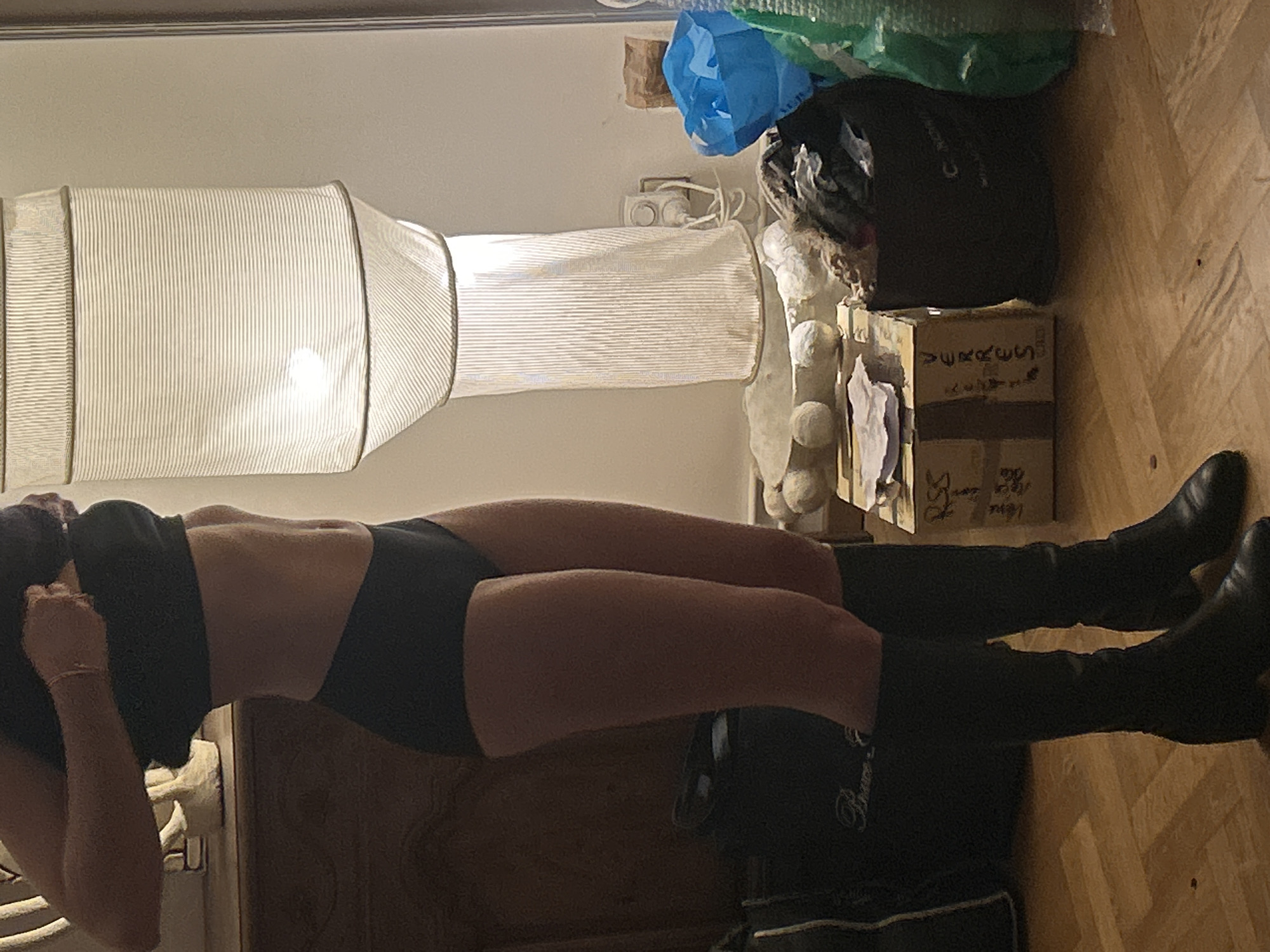 A woman is wearing a black bra, black panties, and knee-high black boots. She is in an indoor setting. A white cylindrical lamp shade hangs from the ceiling. Clutter, including plastic bags, a dark bag, and a cardboard box, is visible in the background. The floor is wooden. The location is unknown.