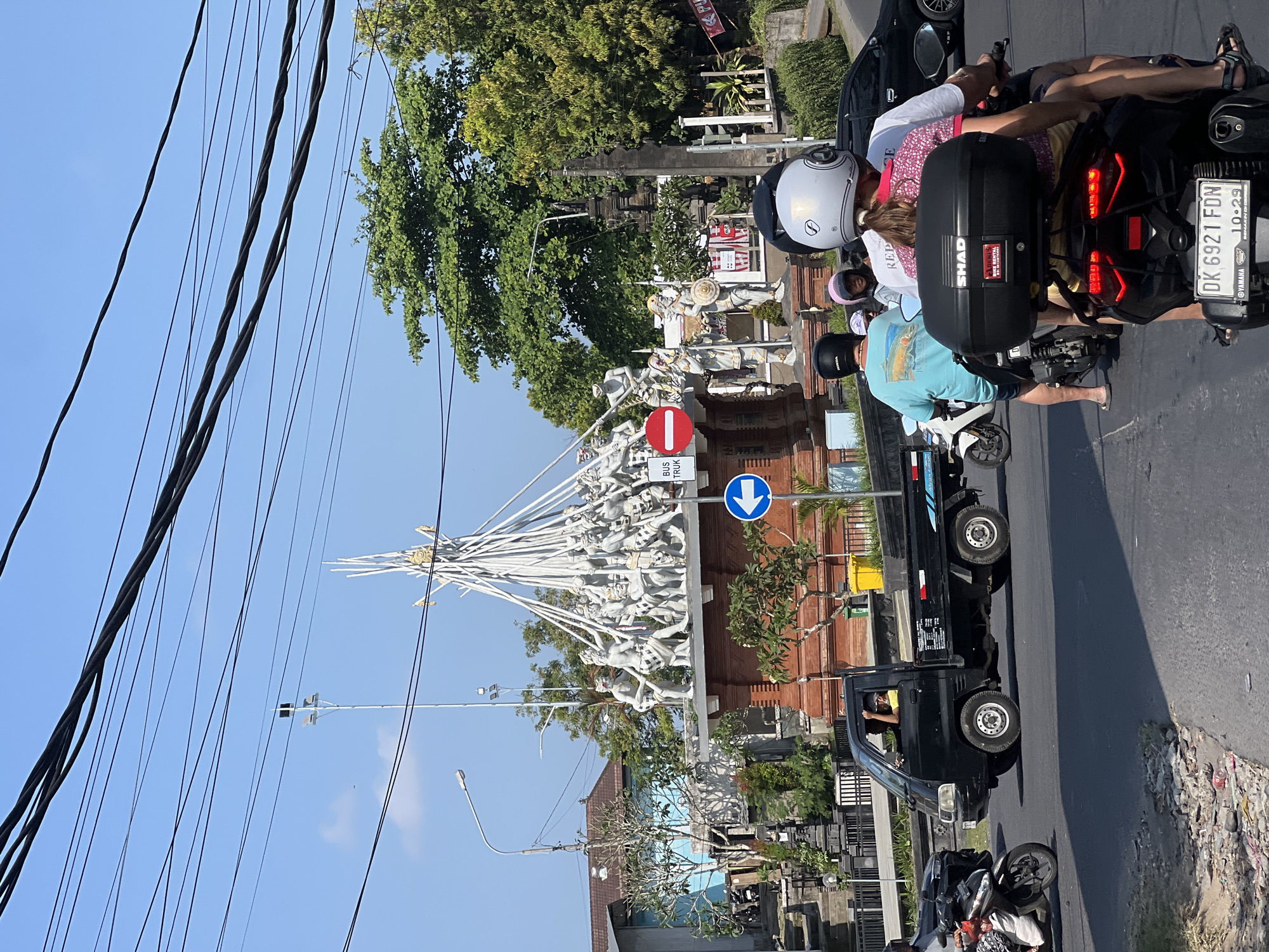 The image captures a street scene in Mengwi, Indonesia, centered around a large monument adorned with numerous white statues depicting figures in what appears to be a dynamic, possibly combat or ritualistic, arrangement. The monument features a tiered, reddish-brown base and is topped with an intricate structure of white poles that fan outwards, supporting the clustered statues. Traffic flows around the monument, with several motorcycles and a black pickup truck visible on the asphalt road.
On the motorcycles, people are wearing helmets, and some are carrying passengers. A man in a yellow shirt is driving the black pickup truck, with his hand raised. The environment is characterized by a clear blue sky, lush green trees bordering the road, and overhead power lines. Buildings with traditional architecture, some with red tiled roofs, are present in the background. Traffic signs, including a blue circular sign with a white arrow indicating a left turn and a red and white circular sign prohibiting something, are strategically placed near the monument. The overall impression is of a busy intersection in a tropical urban setting.