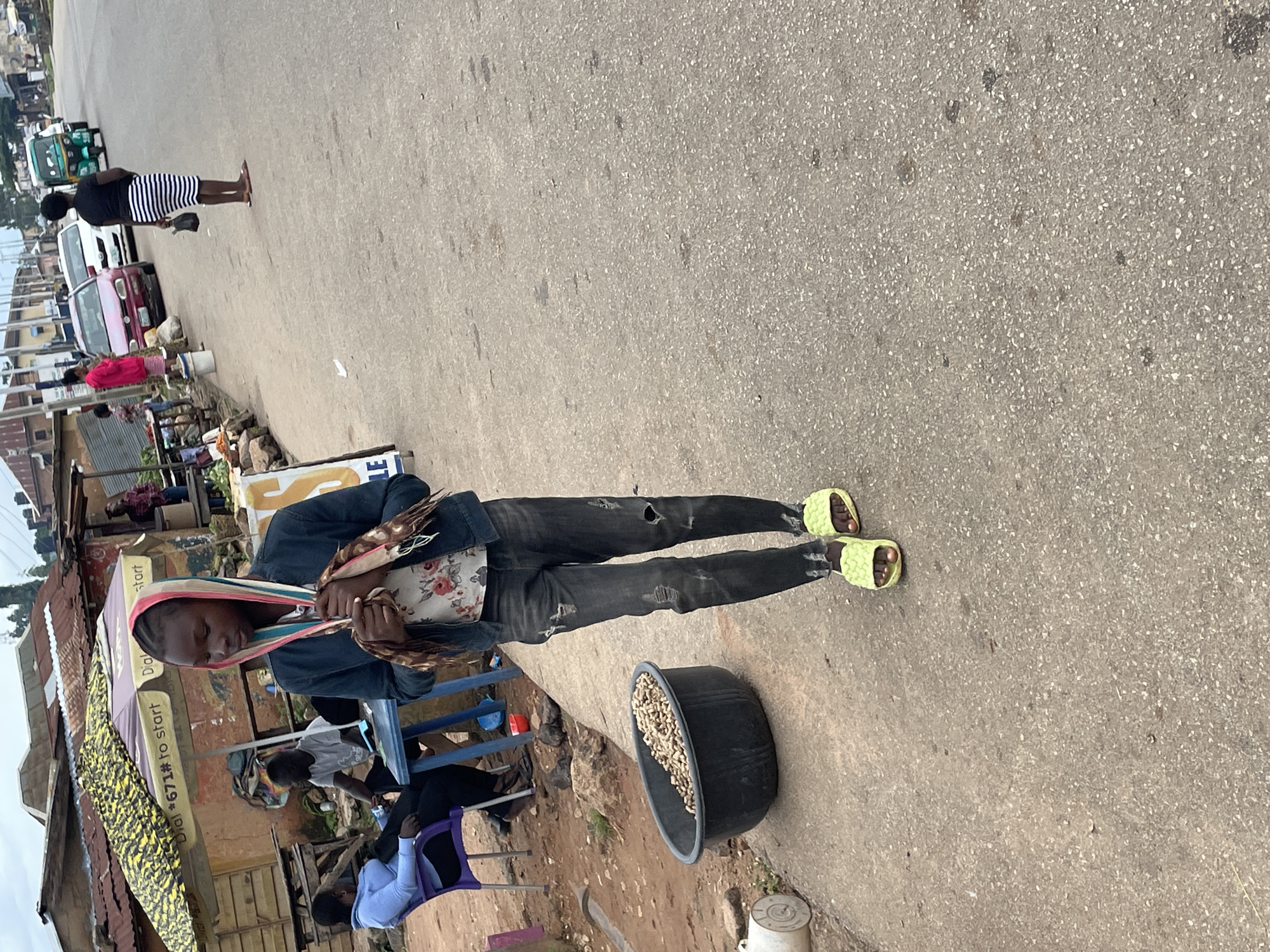 The image shows a street scene in Jos, Nigeria. In the foreground, a young person with dark skin is walking towards the viewer, wearing a dark denim jacket over a floral print shirt and ripped black jeans. They have bright yellow slip-on sandals and are holding a patterned scarf. To the left, a large black basin filled with peanuts is visible on the paved ground. Behind the young person, a covered market stall with a yellow and black patterned awning is set up. Two individuals are seated at a blue table under the awning, with one person wearing a light blue top. Further in the background, a woman in a black and white striped skirt and a black top walks away from the camera, carrying a dark handbag. Other people and vehicles, including a tricycle and cars, are visible on the asphalt road in the distance. The environment appears to be an outdoor market or roadside area.