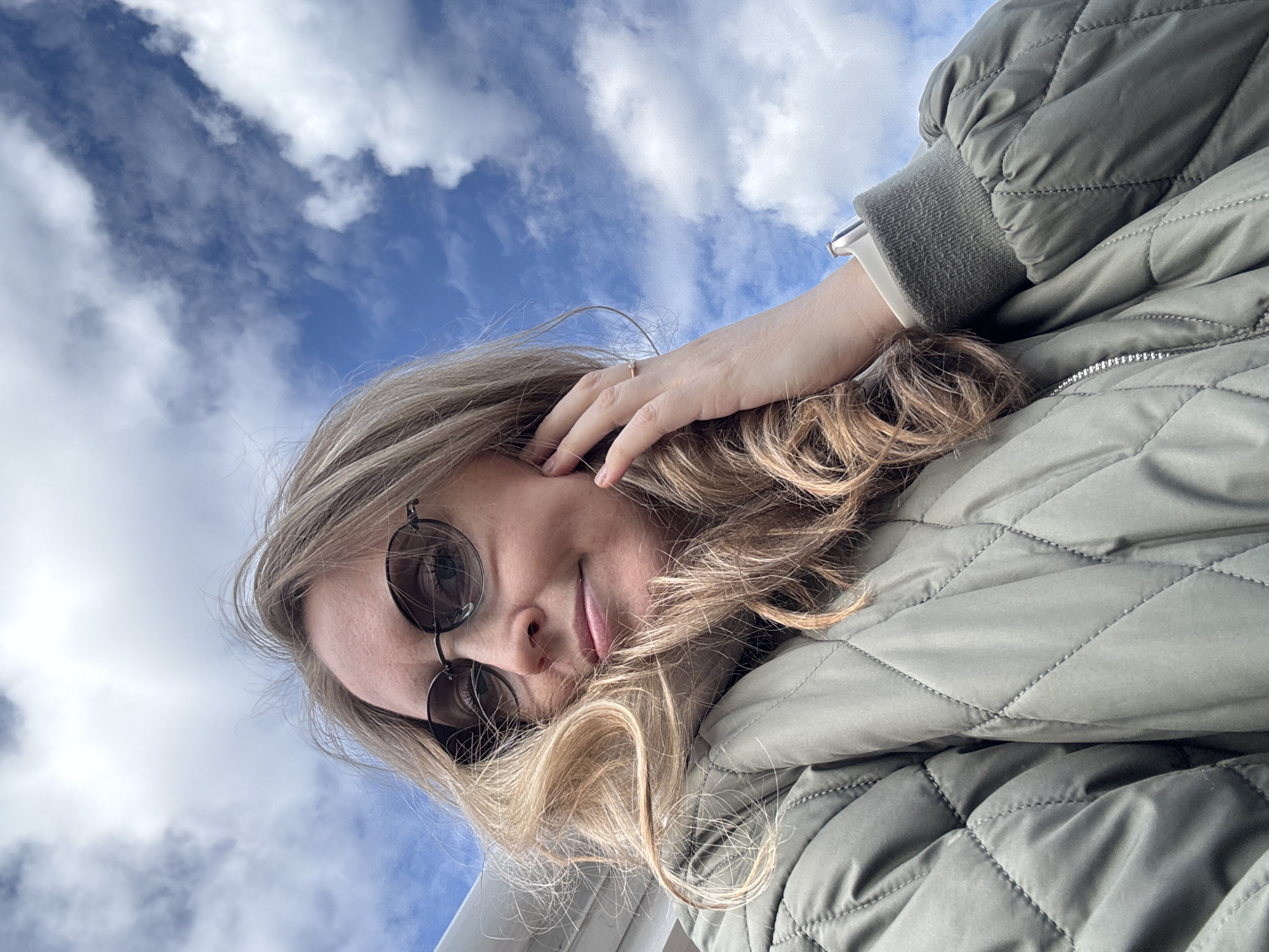 A woman wearing a quilted olive-green jacket and round sunglasses poses against a backdrop of a blue sky with scattered white clouds. She is holding her hand near her head. The location cannot be determined from the visual information.