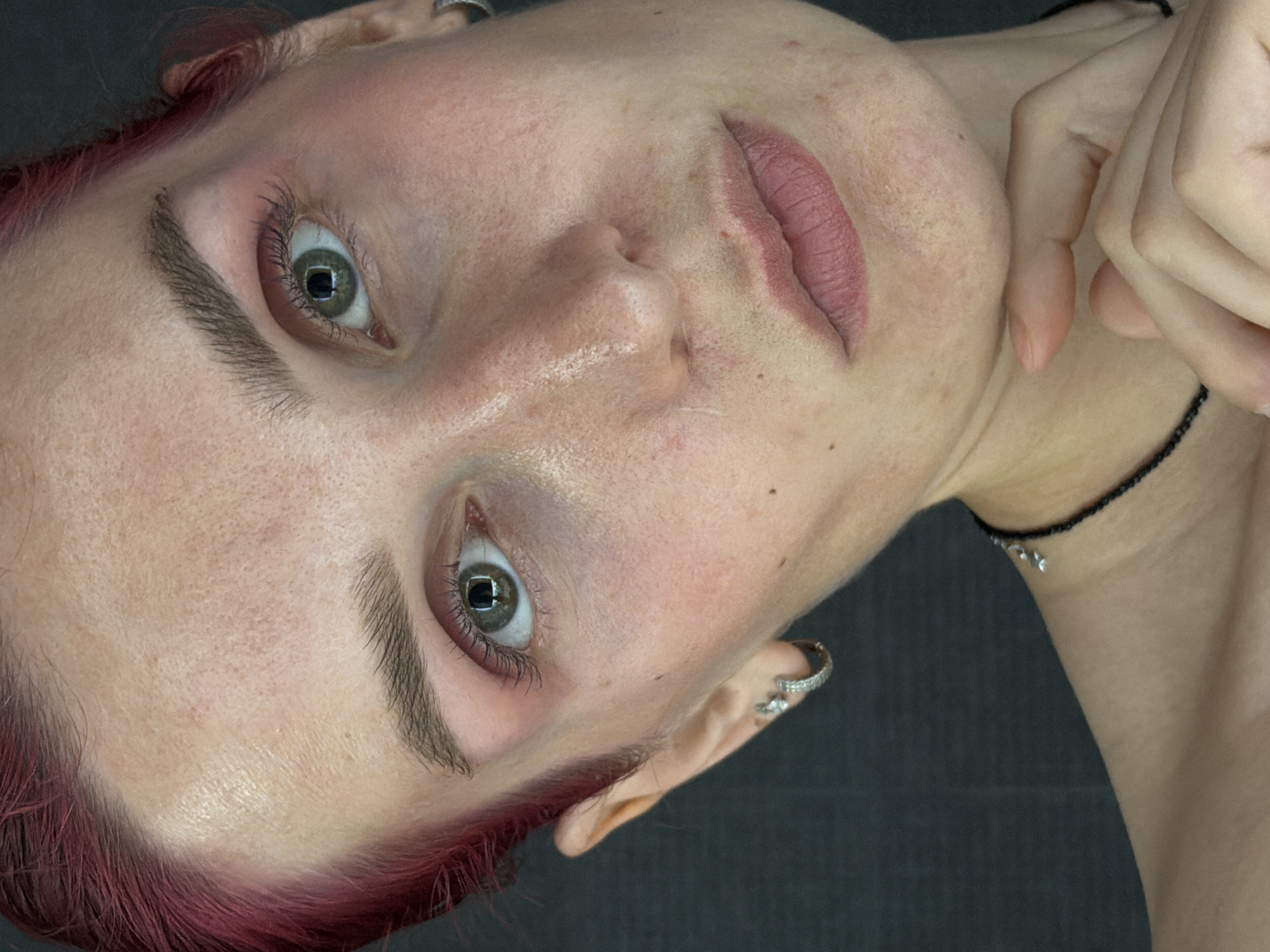 A close-up, portrait-style, high-angle medium shot shows the face of a person with bright pink hair. The person's face is angled slightly to the right, with their chin resting on their right hand. Their eyes are open and looking forward. The person's skin is fair and appears to have freckles and some redness. They are wearing light pink eyeshadow and mascara. A silver hoop earring with small diamonds is visible on their left ear. They are wearing a black beaded necklace with a silver charm. The background is a dark gray, textured surface. The image is well-lit, highlighting the details of the person's face. The image does not contain any information related to the city of Chaikovsky, Russia.