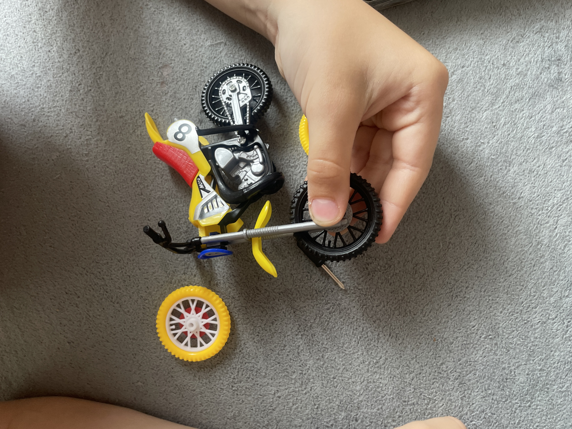 A child's hand holds a toy motorcycle with a yellow, red, and black color scheme against a gray textured surface. A detached yellow wheel with a white and red hub sits near the motorcycle. The motorcycle appears to be partially disassembled. The location is not discernible from the image.