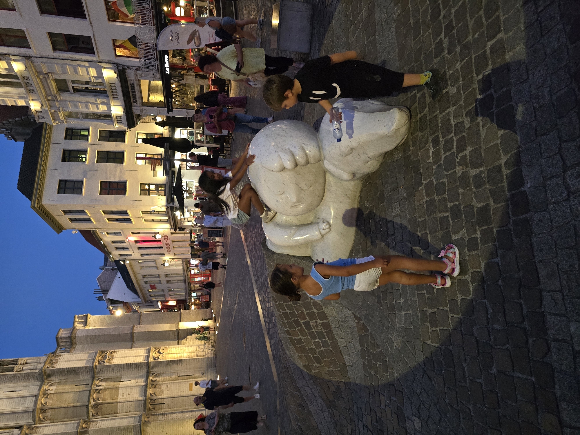 In the evening, in the city of Antwerp, Belgium, a young girl with dark hair, wearing a light blue tank top and white shorts, stands on a cobblestone plaza. To her right, a young boy in a black t-shirt with a smiley face graphic and black pants, holding a water bottle, looks towards her. Behind them, a large, abstract white sculpture, resembling a sleeping animal, is positioned on the plaza. The sculpture appears to be made of polished stone or marble. Further in the background, several adults are visible walking along a street lined with buildings that are illuminated. Some of these buildings have awnings and signs that suggest they are shops or restaurants. The sky is a deep blue, indicating dusk or early evening. The overall atmosphere is one of a public square in a European city at the end of the day.